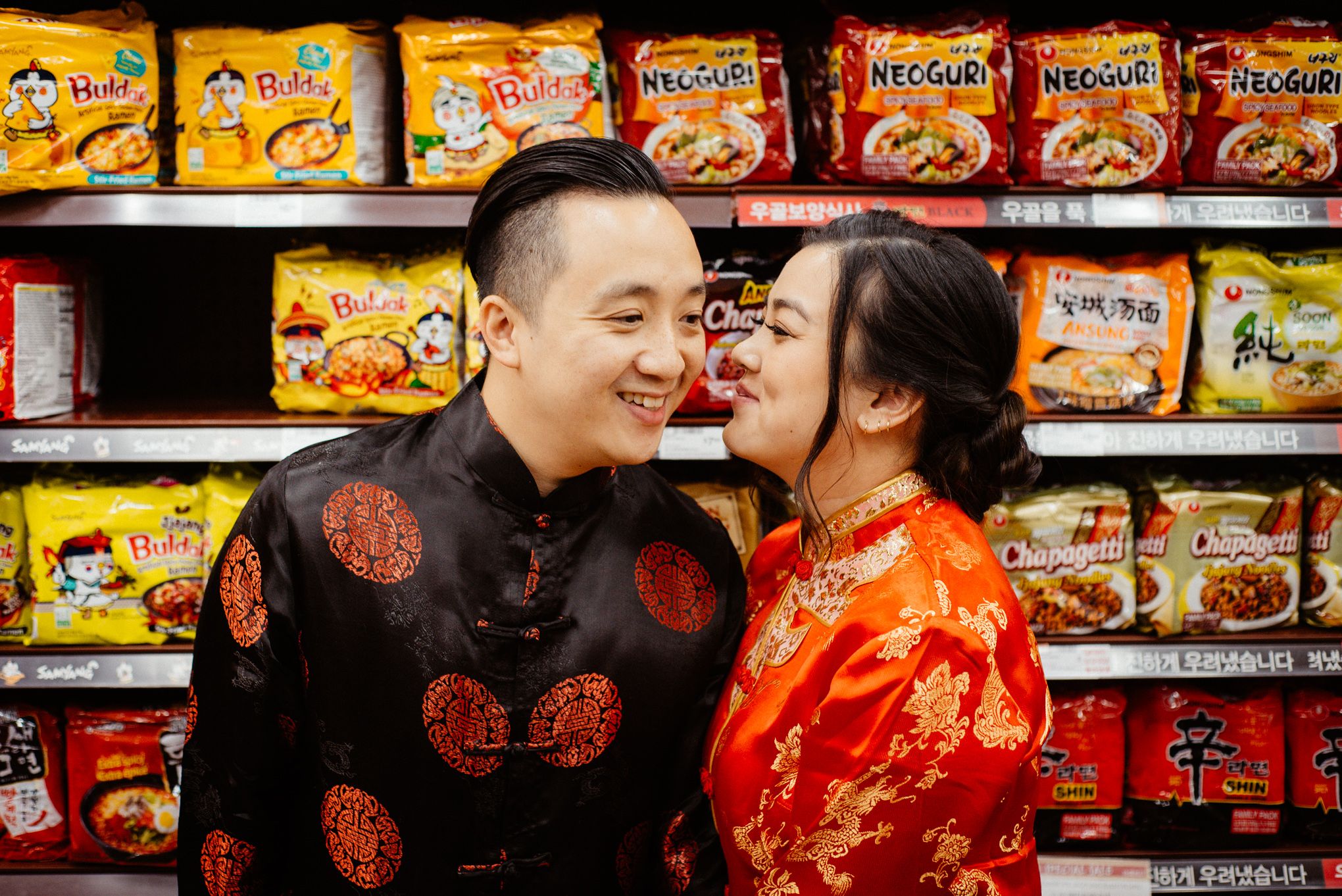 Couple poses near instant noodle shelf in grocery store wearing traditional red attire.