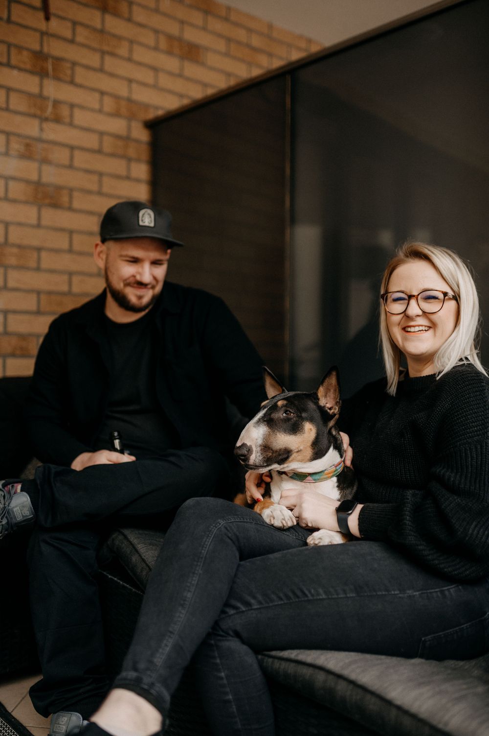 Smiling couple sits on a sofa with their bull terrier dog in a modern home.