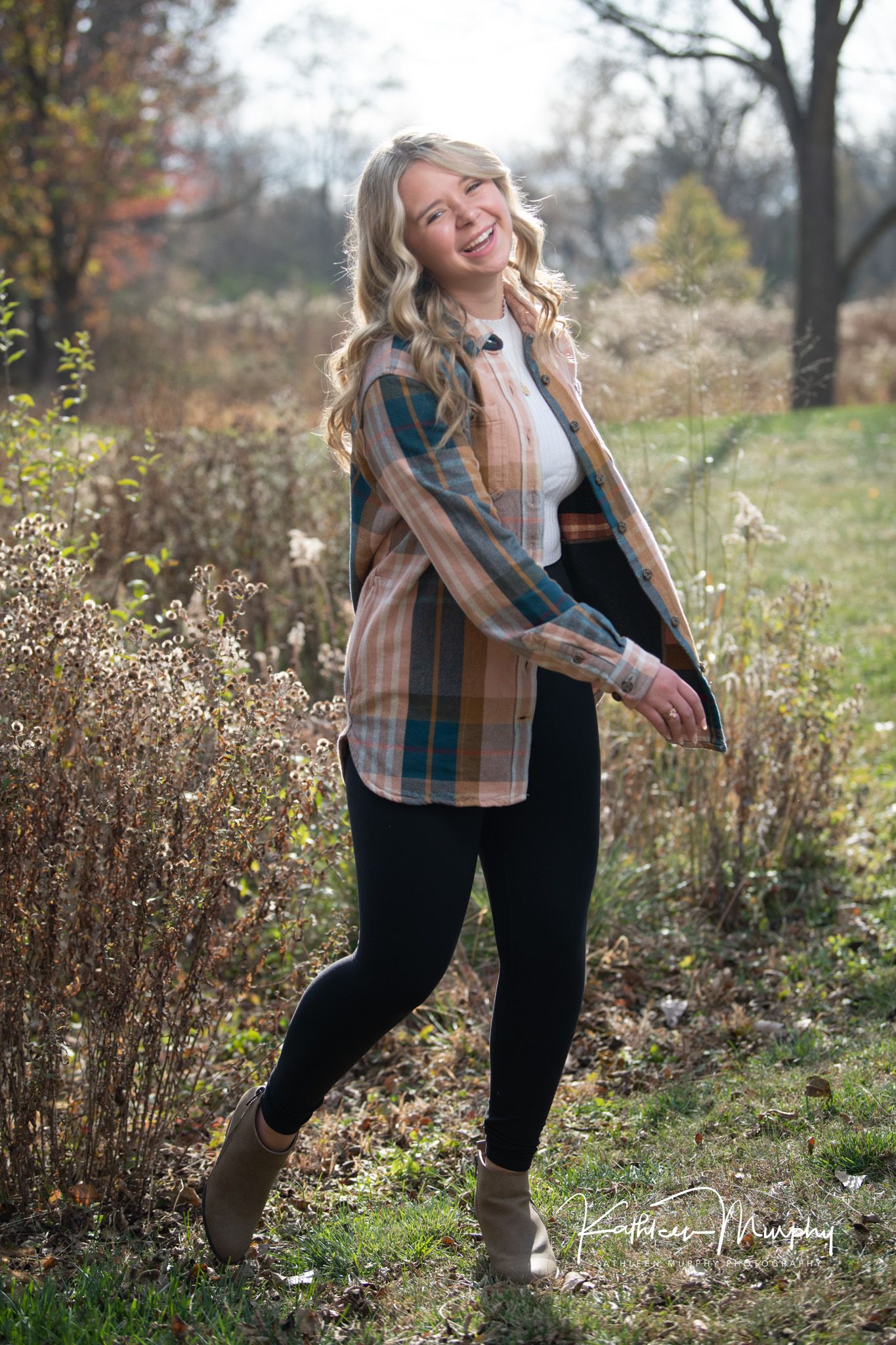 Natural outdoor portrait in plaid shirt and black pants, posing playfully among fall foliage.