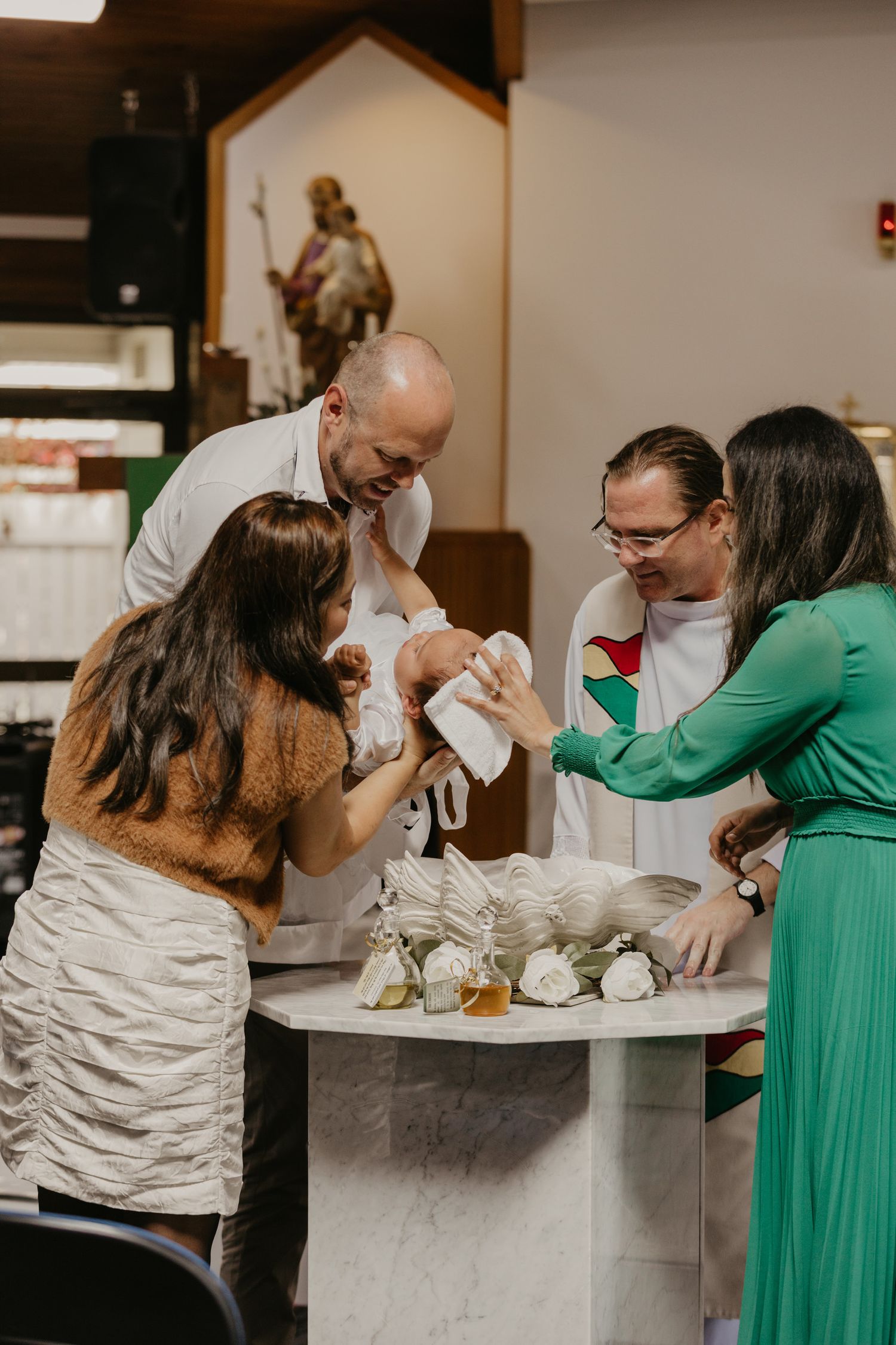 Group gathers around table for religious baptism ceremony with priest in church setting.