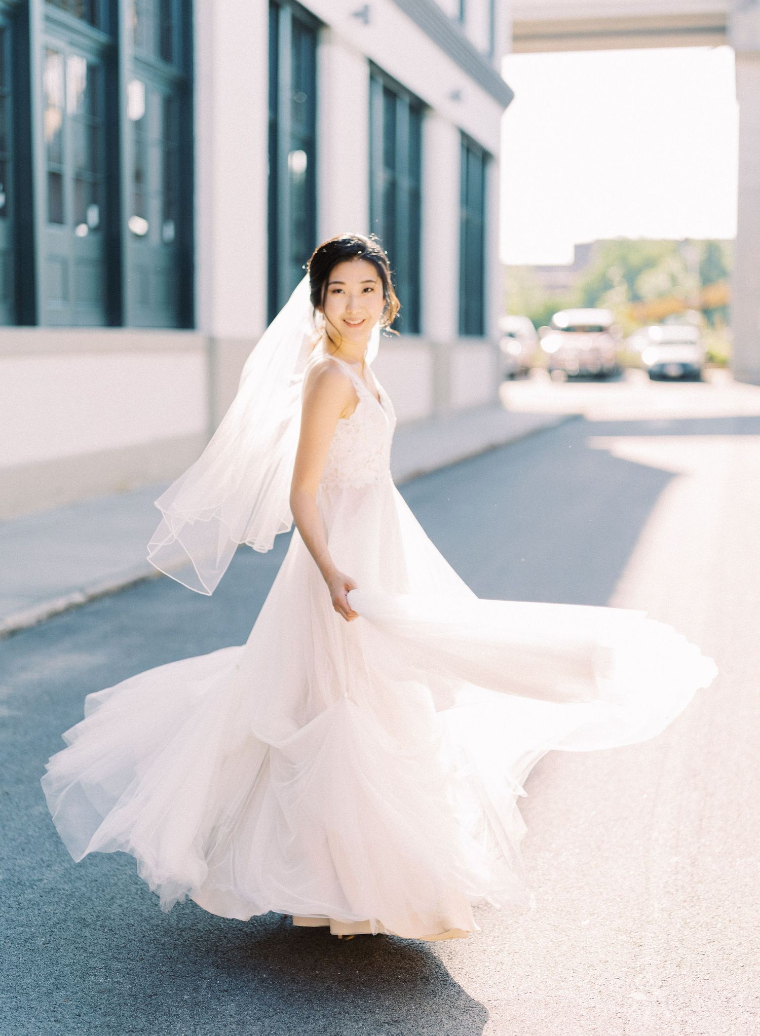 Bride in flowing white dress twirling with veil on urban sidewalk.