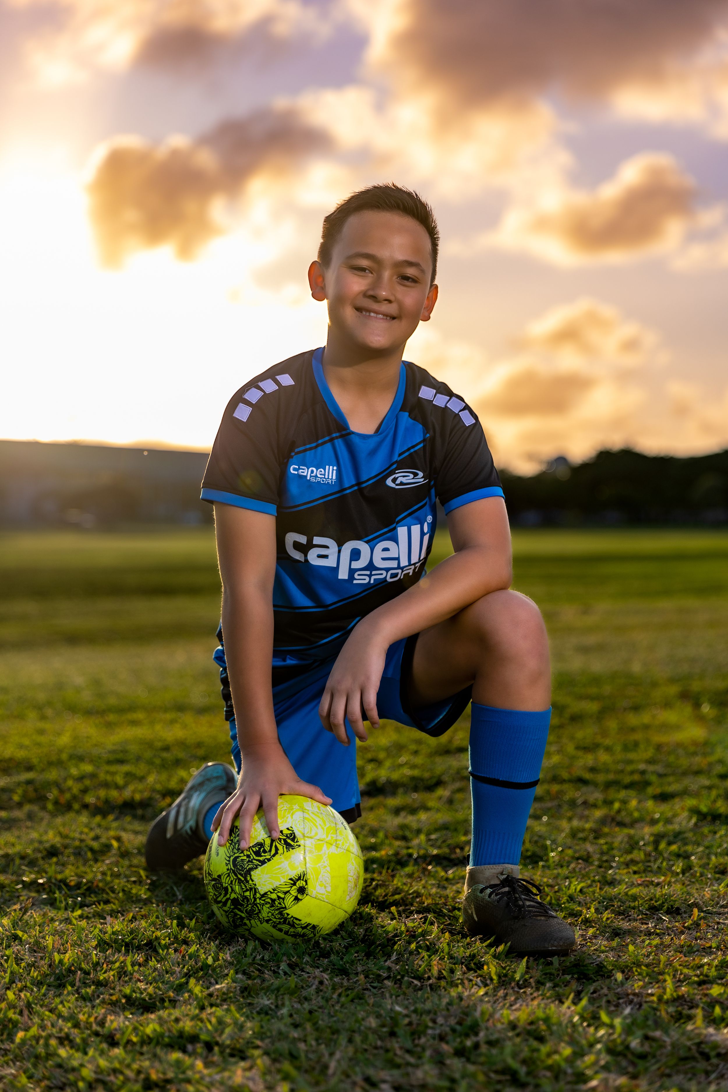 Soccer player in blue Capelli uniform poses with ball during golden sunset.