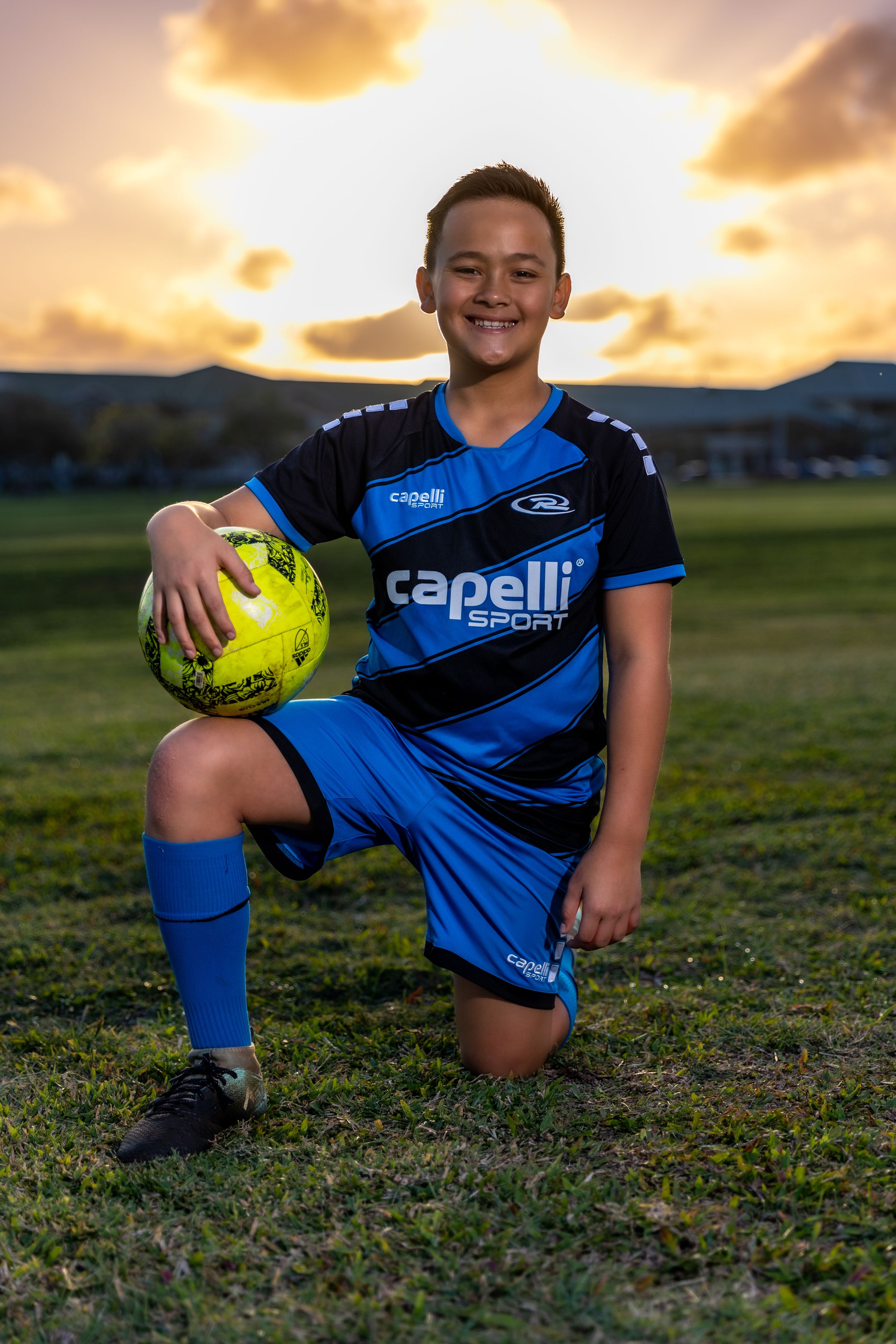 Young athlete in blue Capelli jersey resting with soccer ball at sunset.