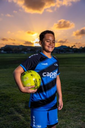 Soccer player holding yellow ball while wearing blue Capelli uniform at dusk.