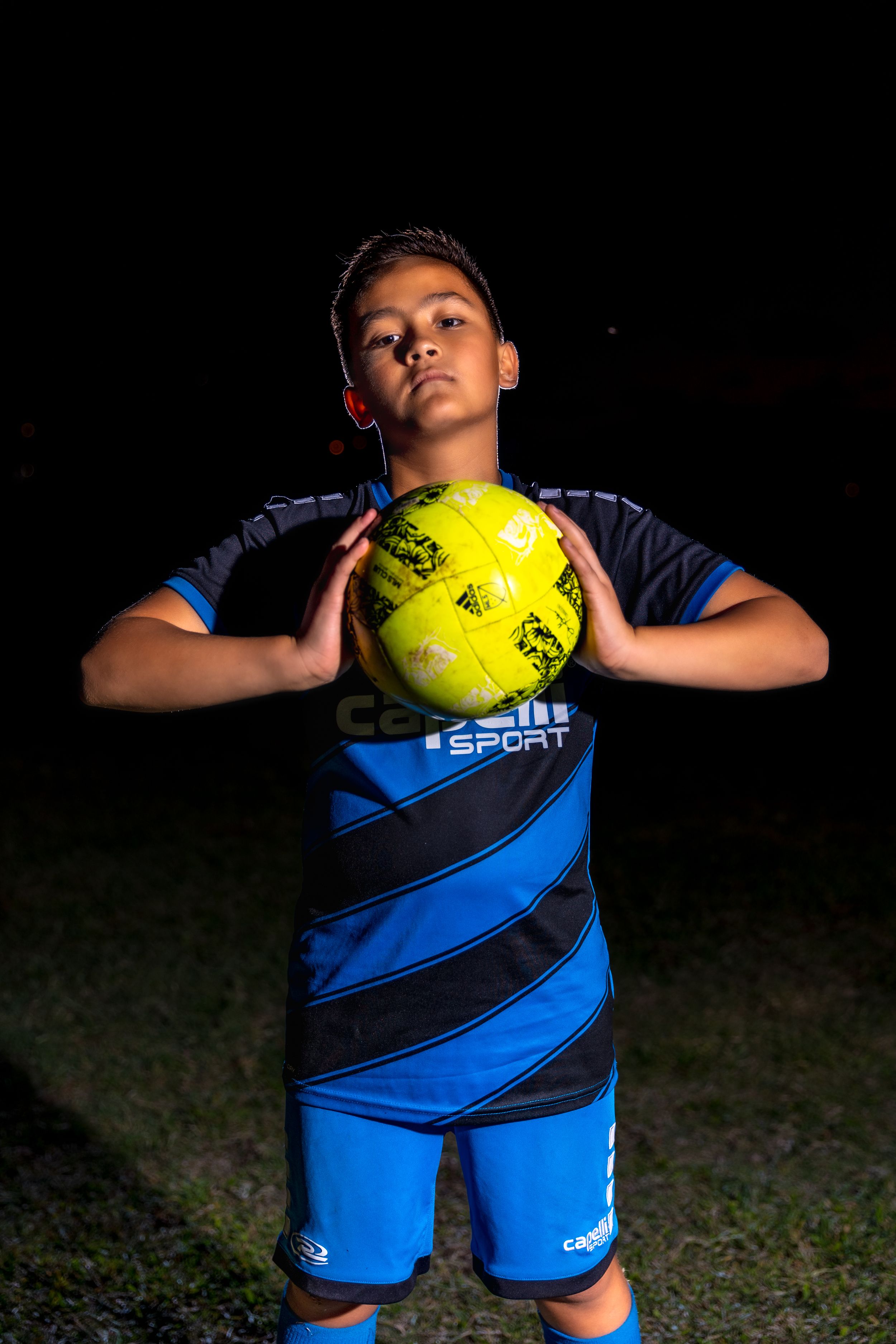 Soccer player in blue uniform demonstrates ball control techniques with a yellow ball against dark background.