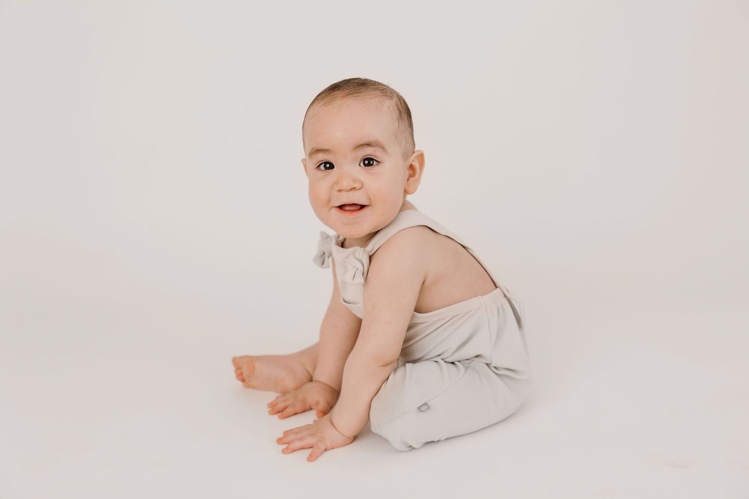 Baby in light gray outfit sits on white studio background.