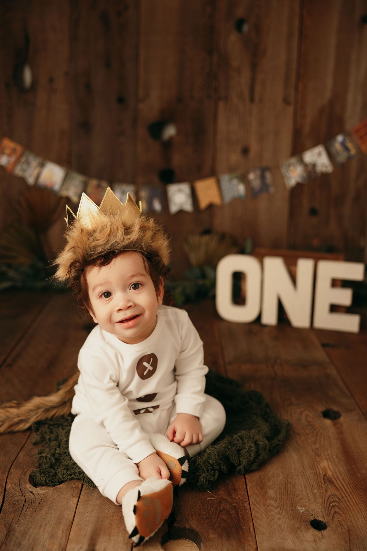 Baby in white outfit with crown poses by ONE sign and banner on wooden background.