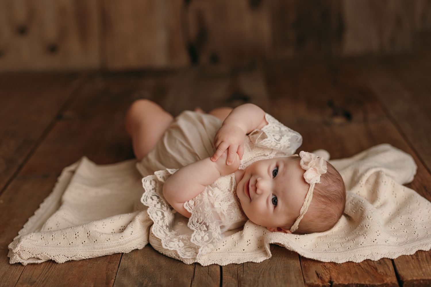 Baby in vintage lace dress lying on wooden floor with cream fabric.