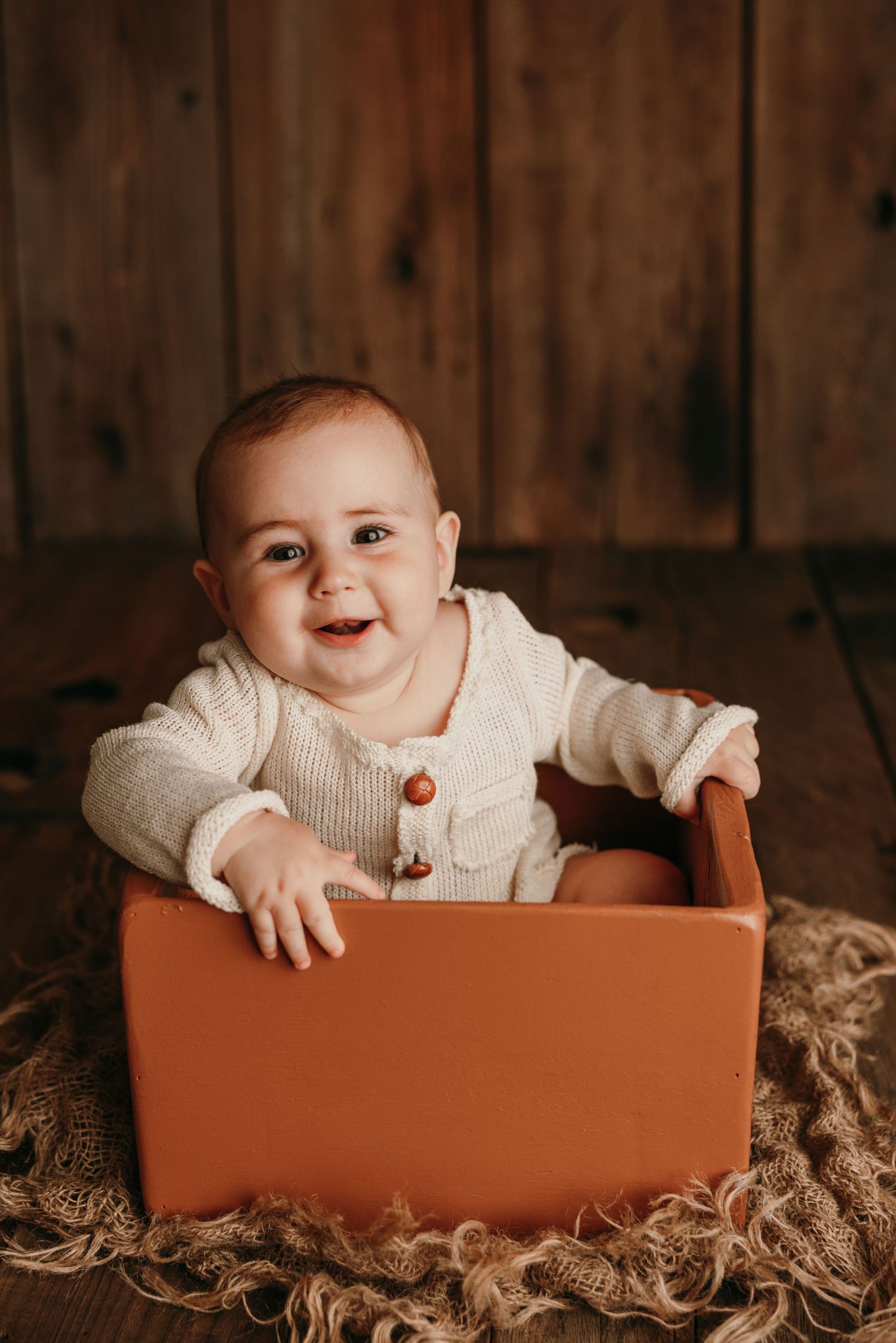 Baby sitting in terra cotta colored box wearing cream knit sweater against wooden backdrop.