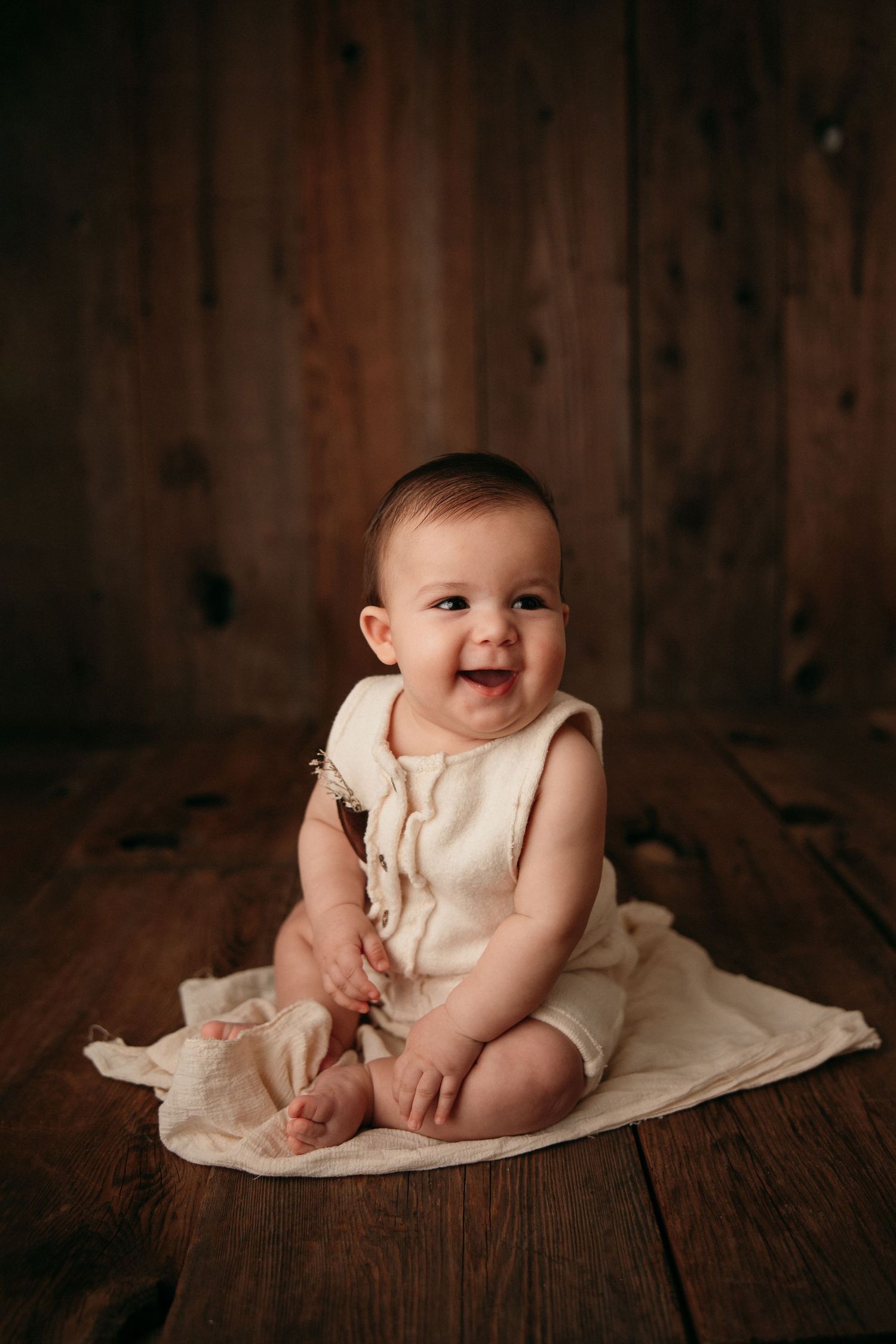 Baby in cream colored outfit sitting on wooden floor with joyful expression.
