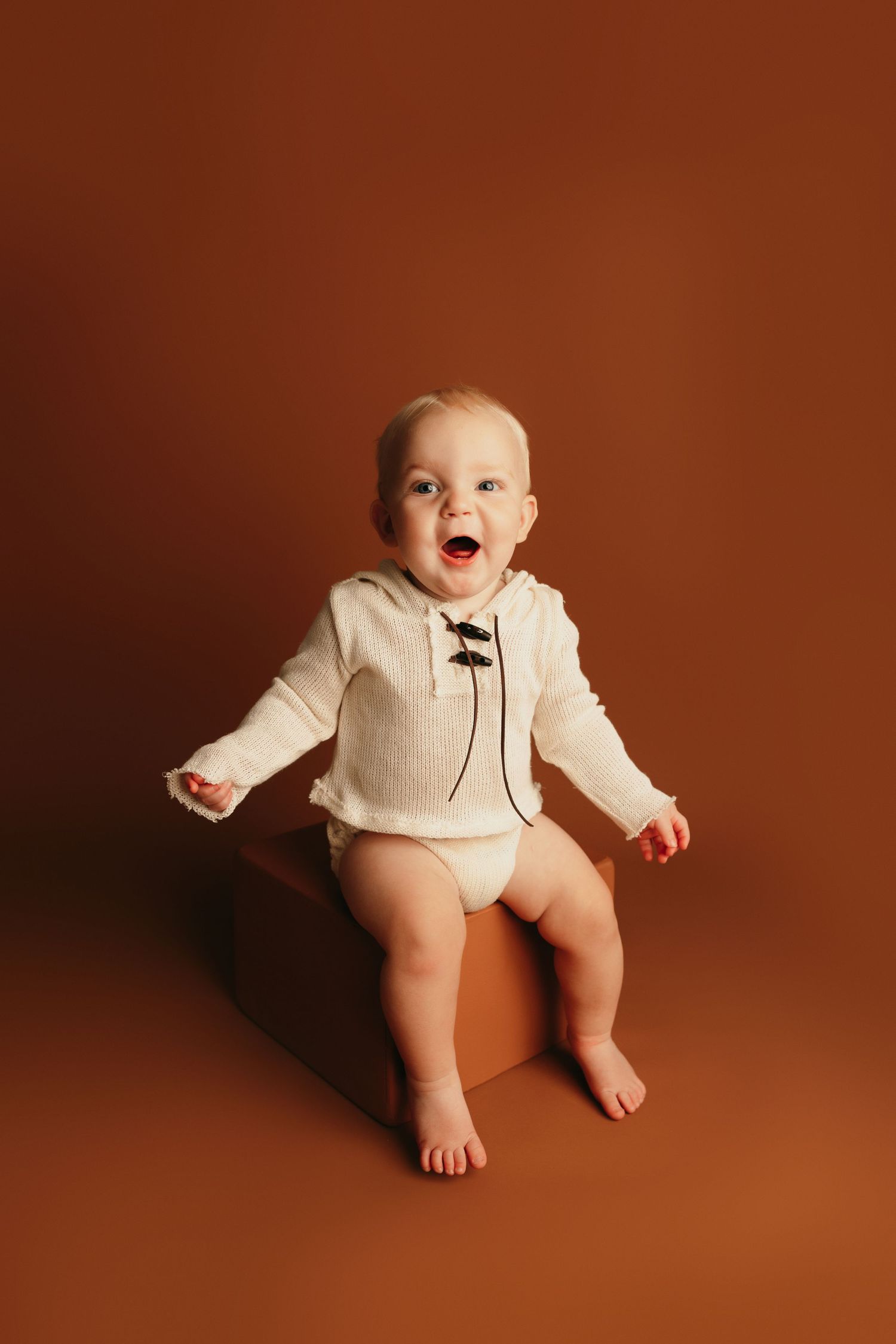Baby in cream sweater shows excitement against warm brown studio background.