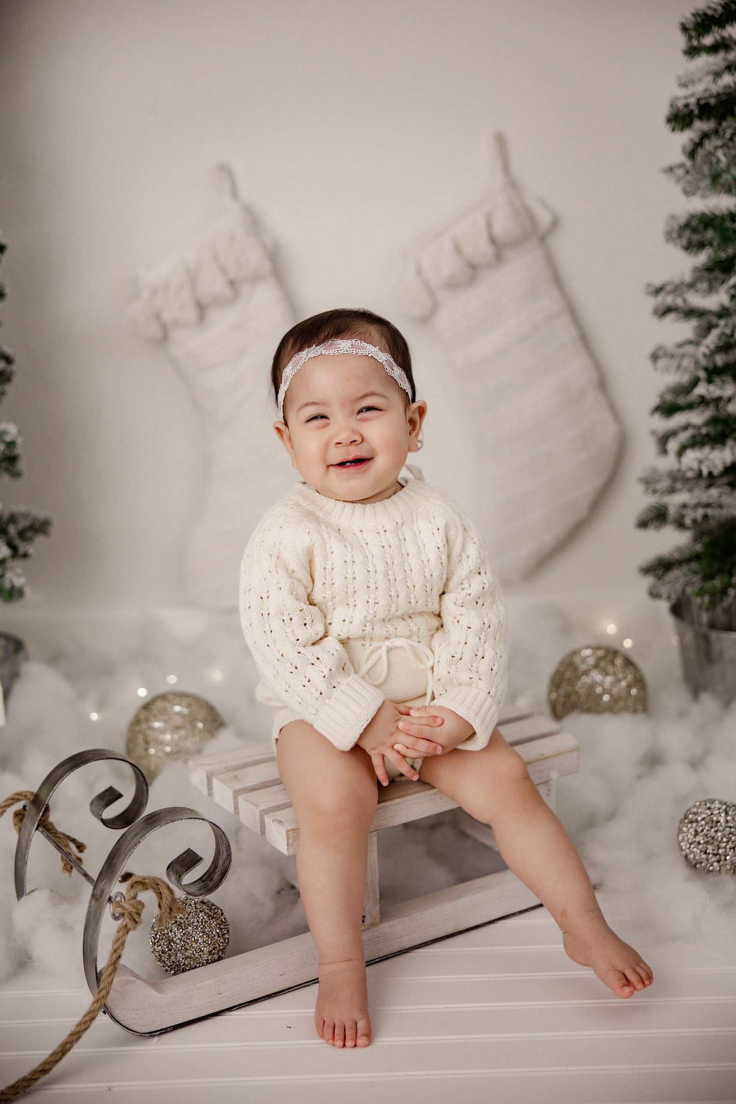 Baby wearing cream knit sweater sitting on white bench with holiday decorations and stockings.