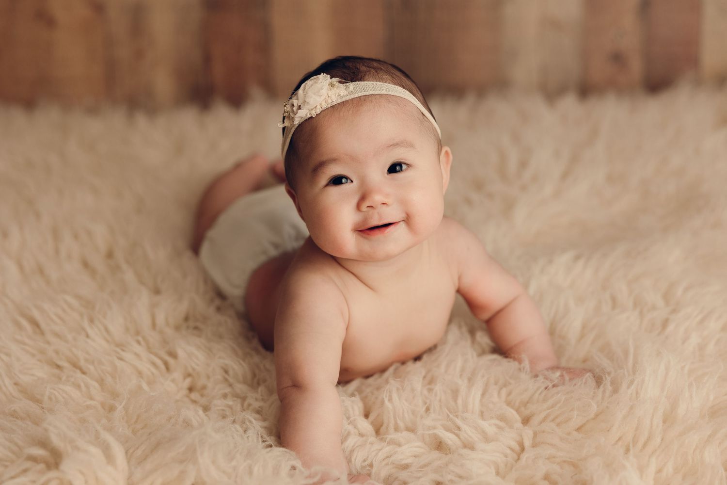 Baby lying on soft beige fur rug wearing a lace headband during professional photoshoot.