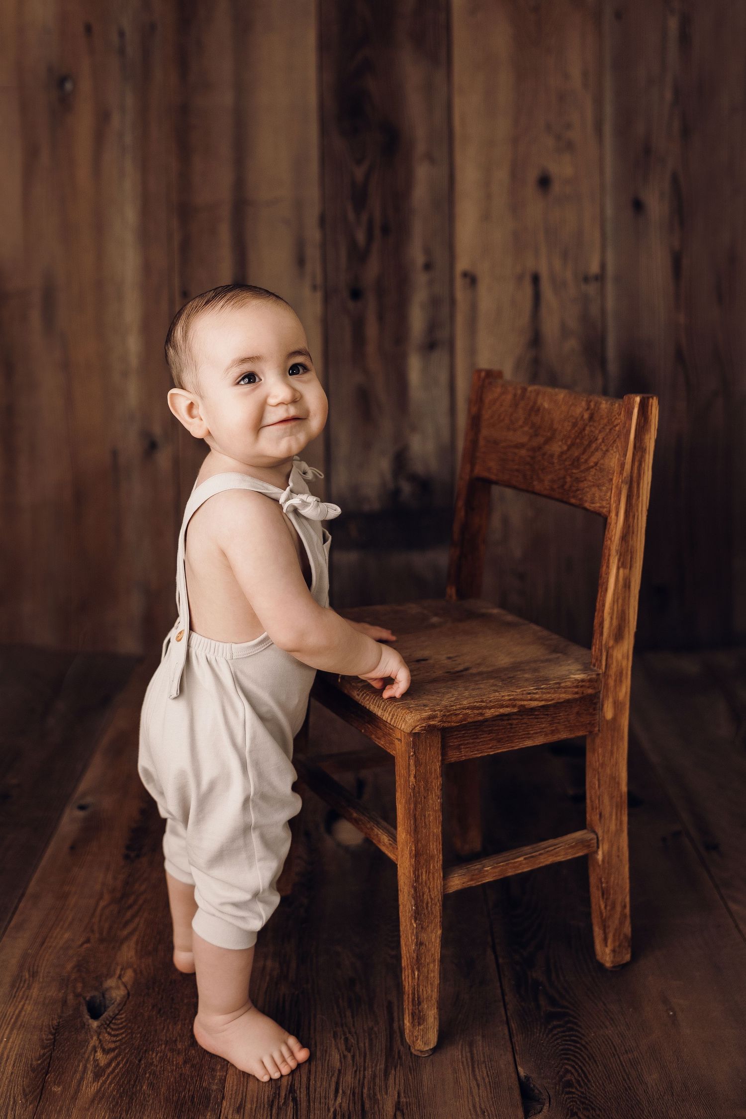 Baby in light colored romper stands beside wooden chair against rustic wall.