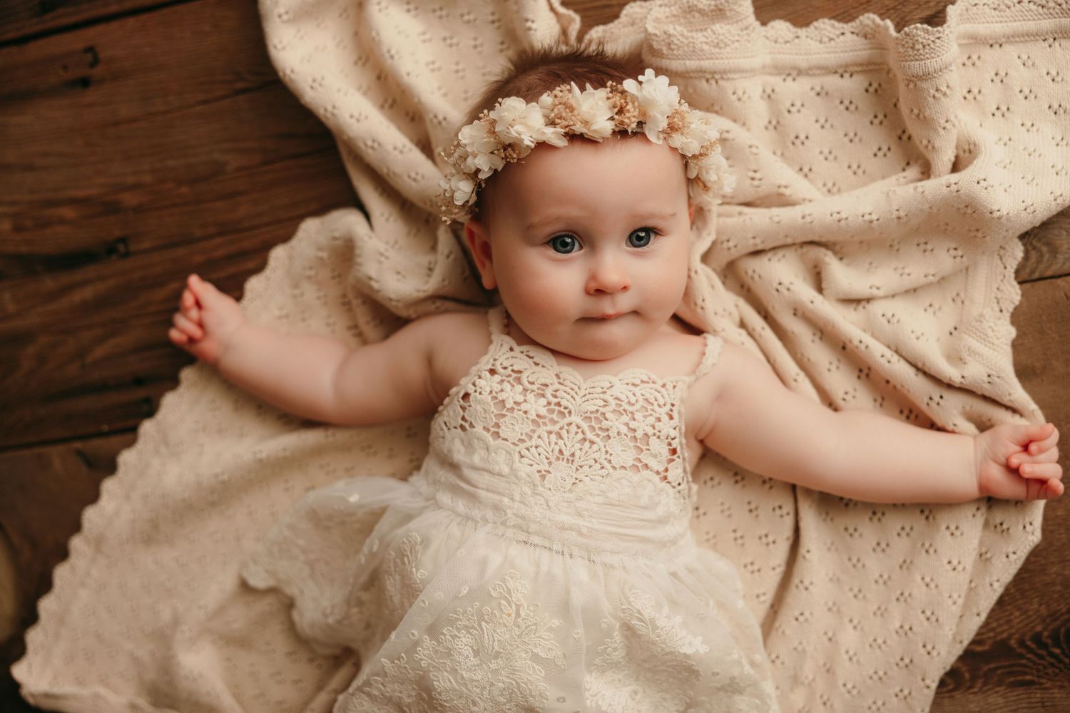 Baby wearing lace dress and floral crown lies on cream fabric against wooden background.