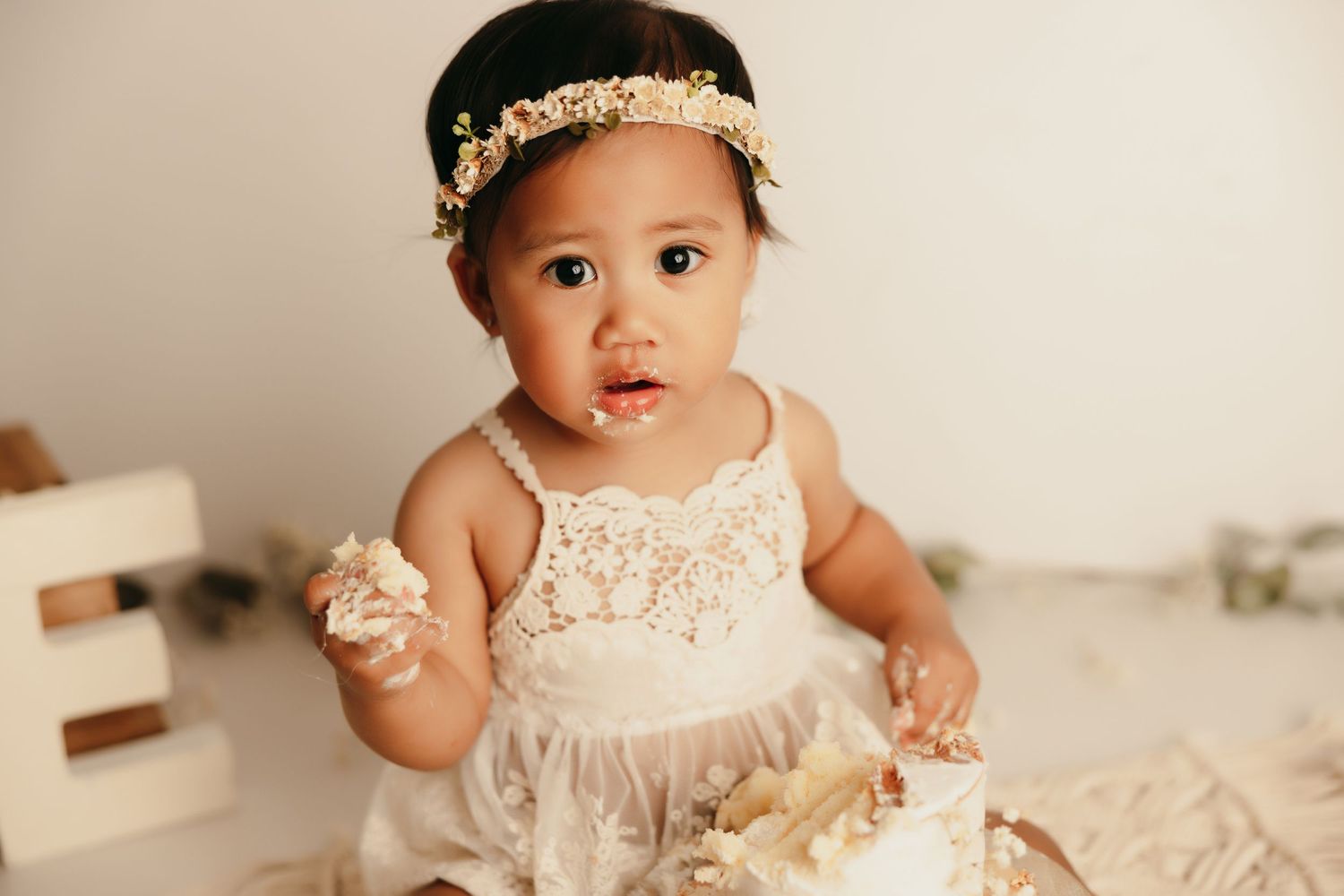 Baby in white lace dress with gold headband sitting with cake during first birthday photoshoot.