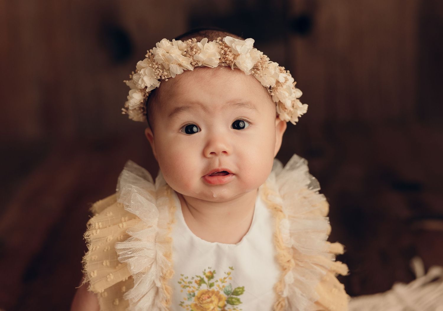 Baby wearing a cream floral dress and lace headband with delicate ruffled details against dark wood background.