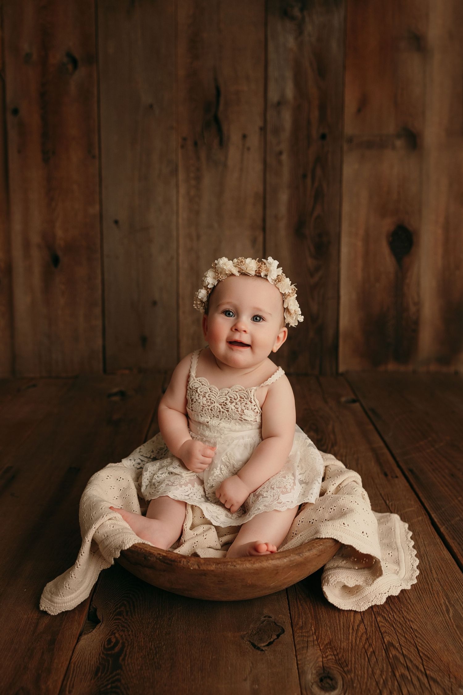 Joyful baby in lace dress with flower crown sitting in wooden bowl.