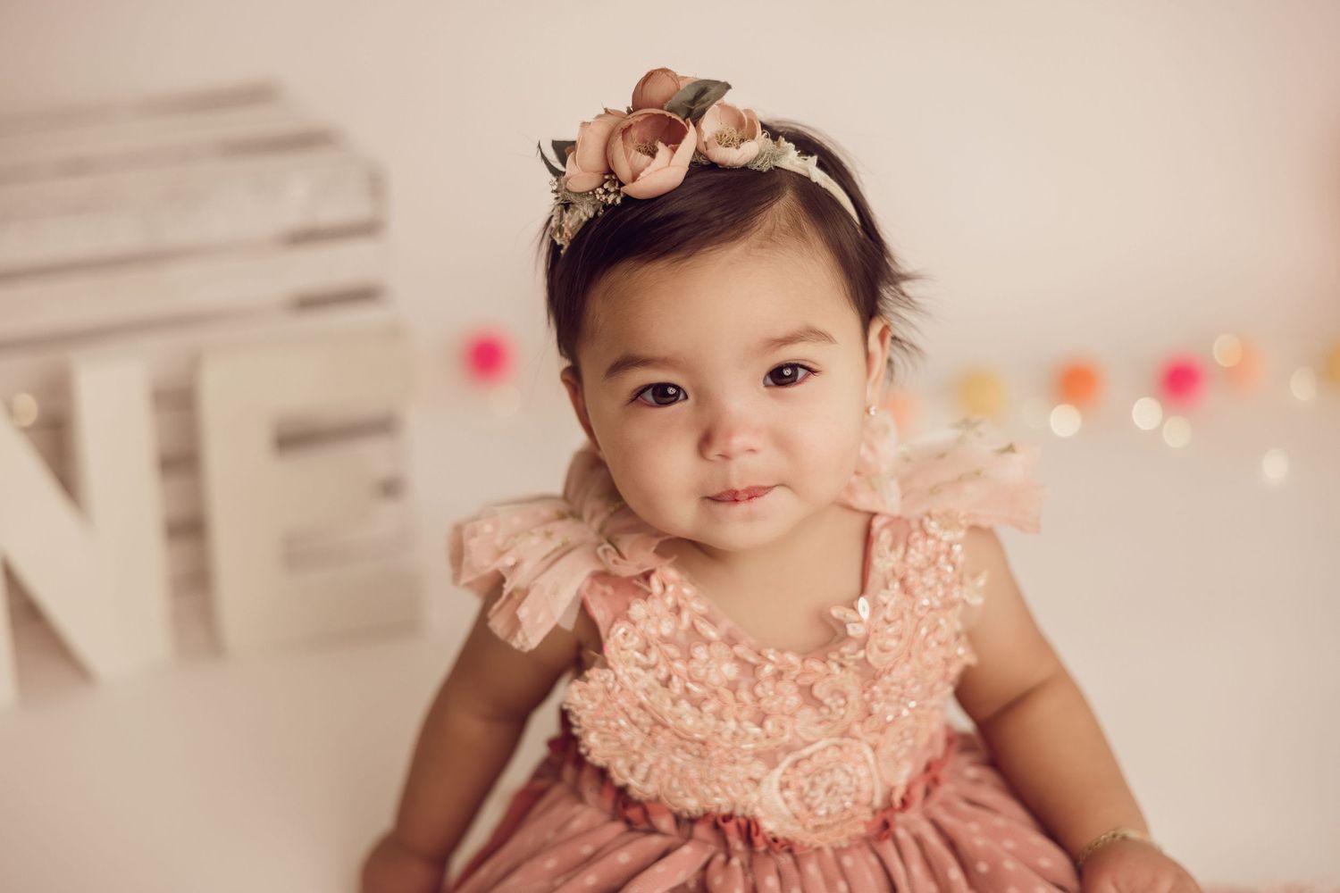 Baby in pink lace dress with bow headband poses against light background.