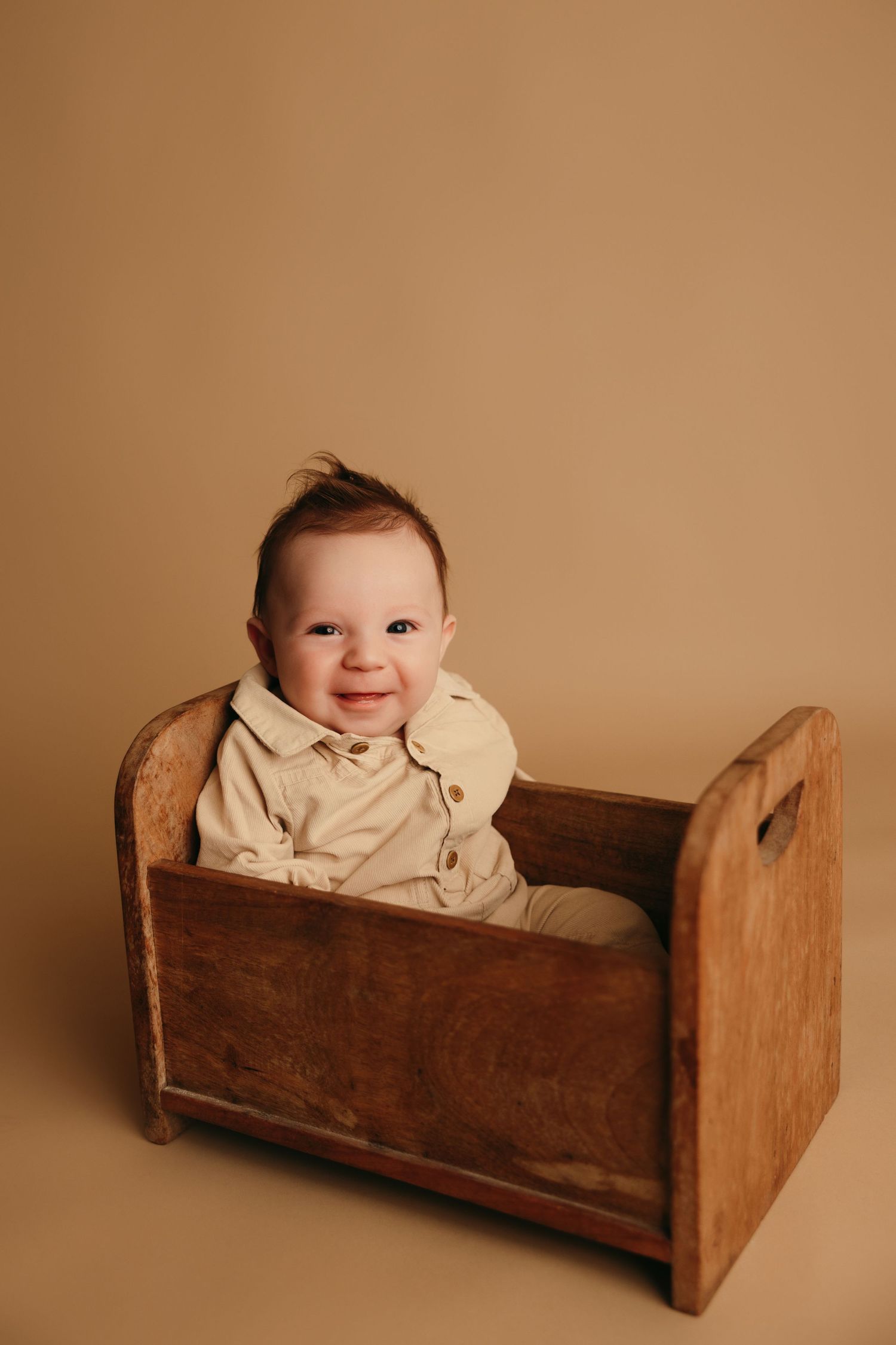 Baby in beige outfit sits in vintage wooden cradle against neutral background.