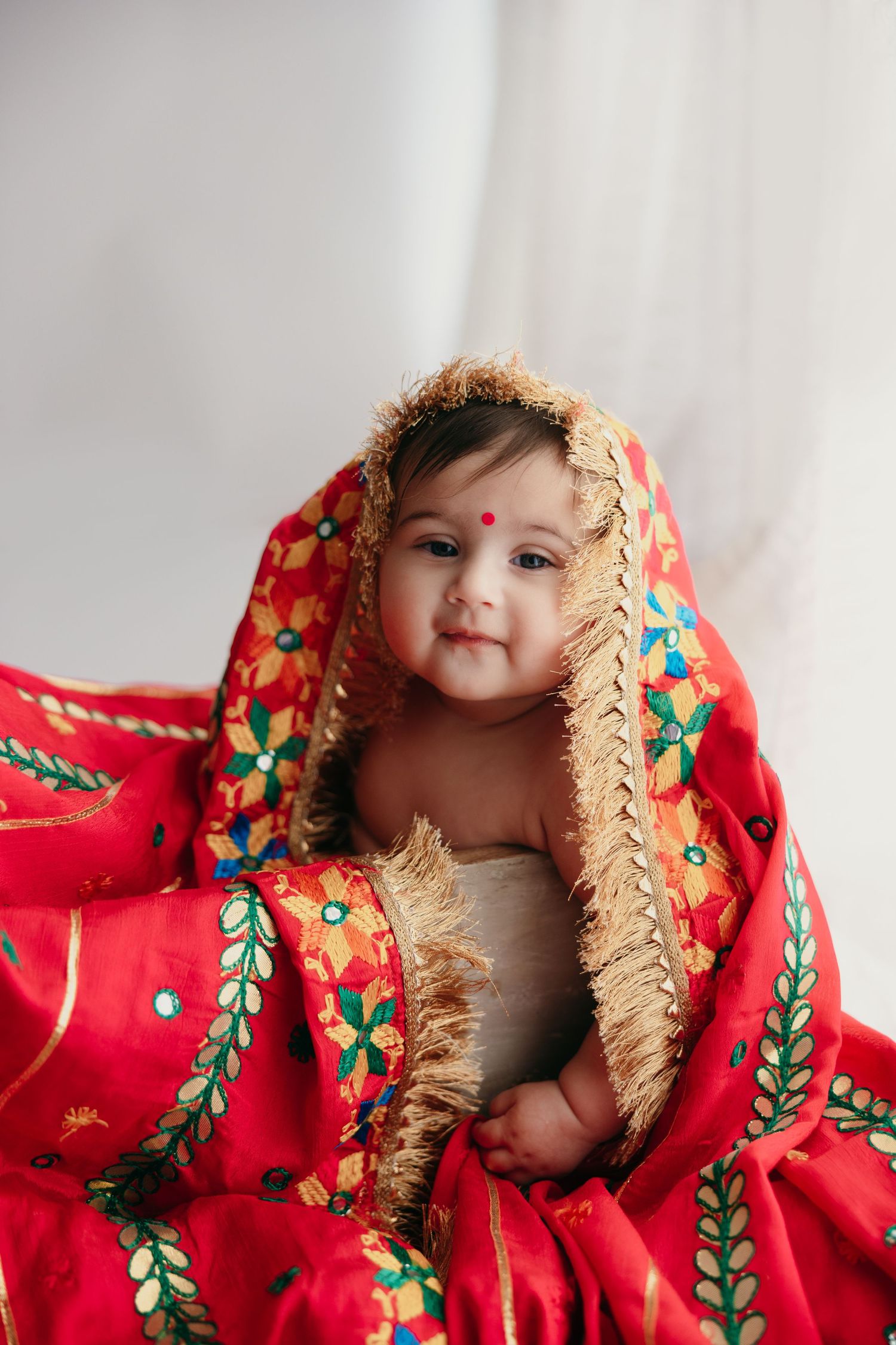 Smiling baby wrapped in vibrant red floral embroidered traditional shawl.