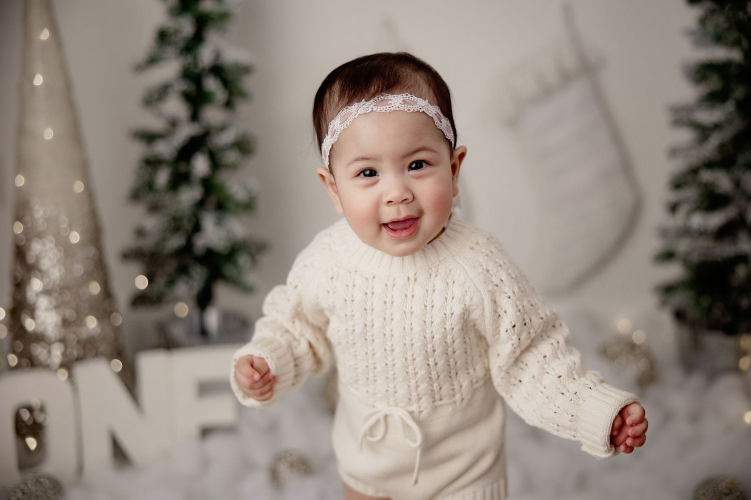 Baby in white knit sweater smiling in winter themed setting with Christmas stockings and trees.