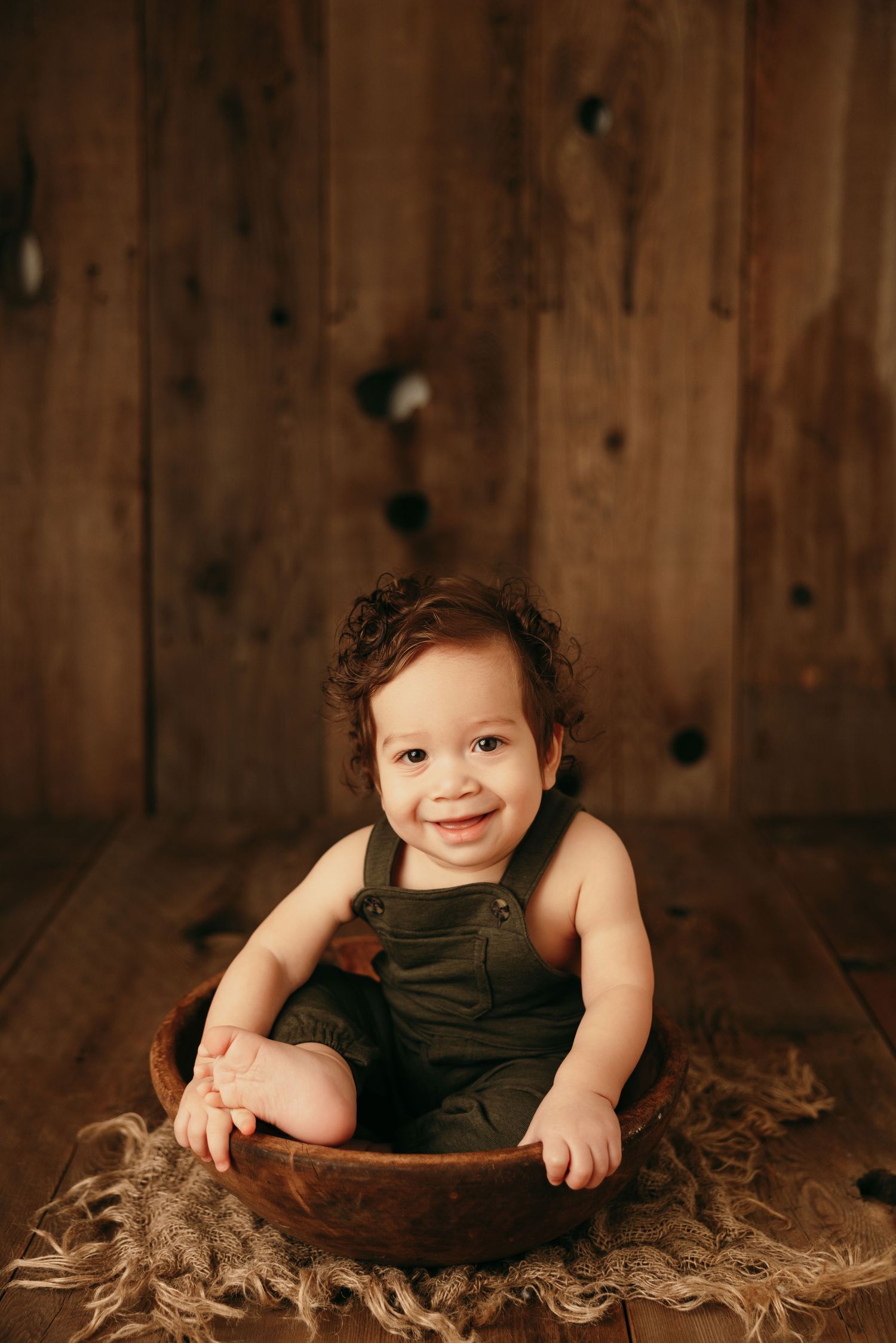 Smiling baby in dark overalls sits in wooden bowl on rustic backdrop.