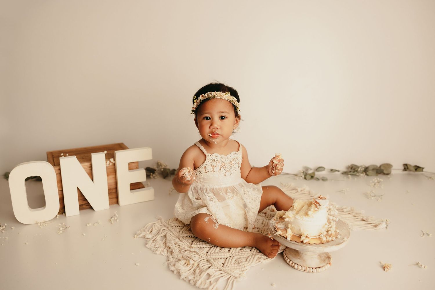 Baby in white dress sitting next to 'ONE' letters and cake during first birthday celebration.