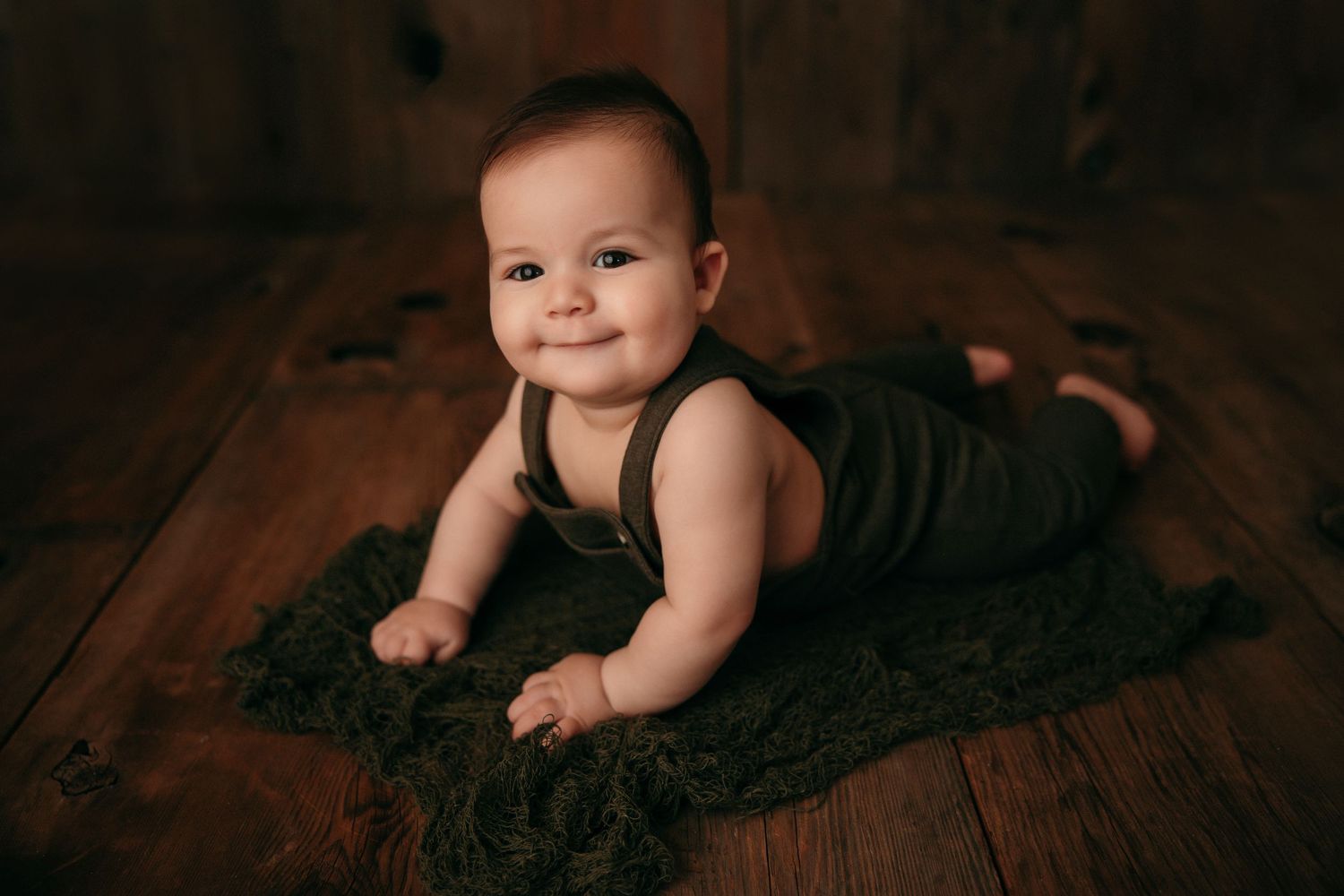 Baby in dark outfit lying on textured blanket against rustic wooden floor background.