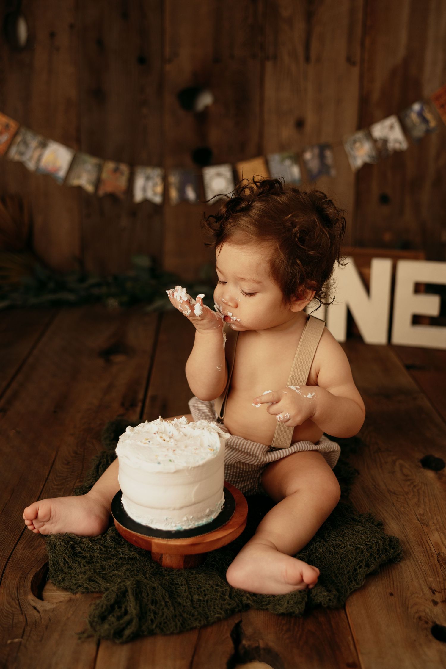 Baby in overalls enjoys first birthday cake smash on wooden floor with ONE sign.