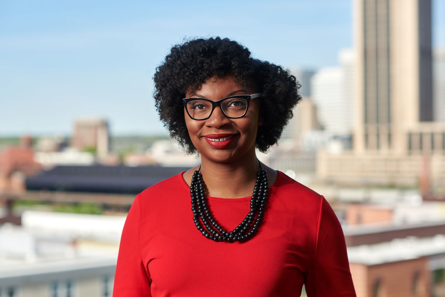 Professional headshot of person wearing glasses and red outfit against urban backdrop.