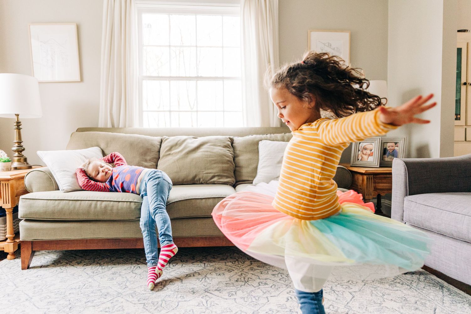 A young girl is laying on the couch and watching her twin sister twirl in a tutu