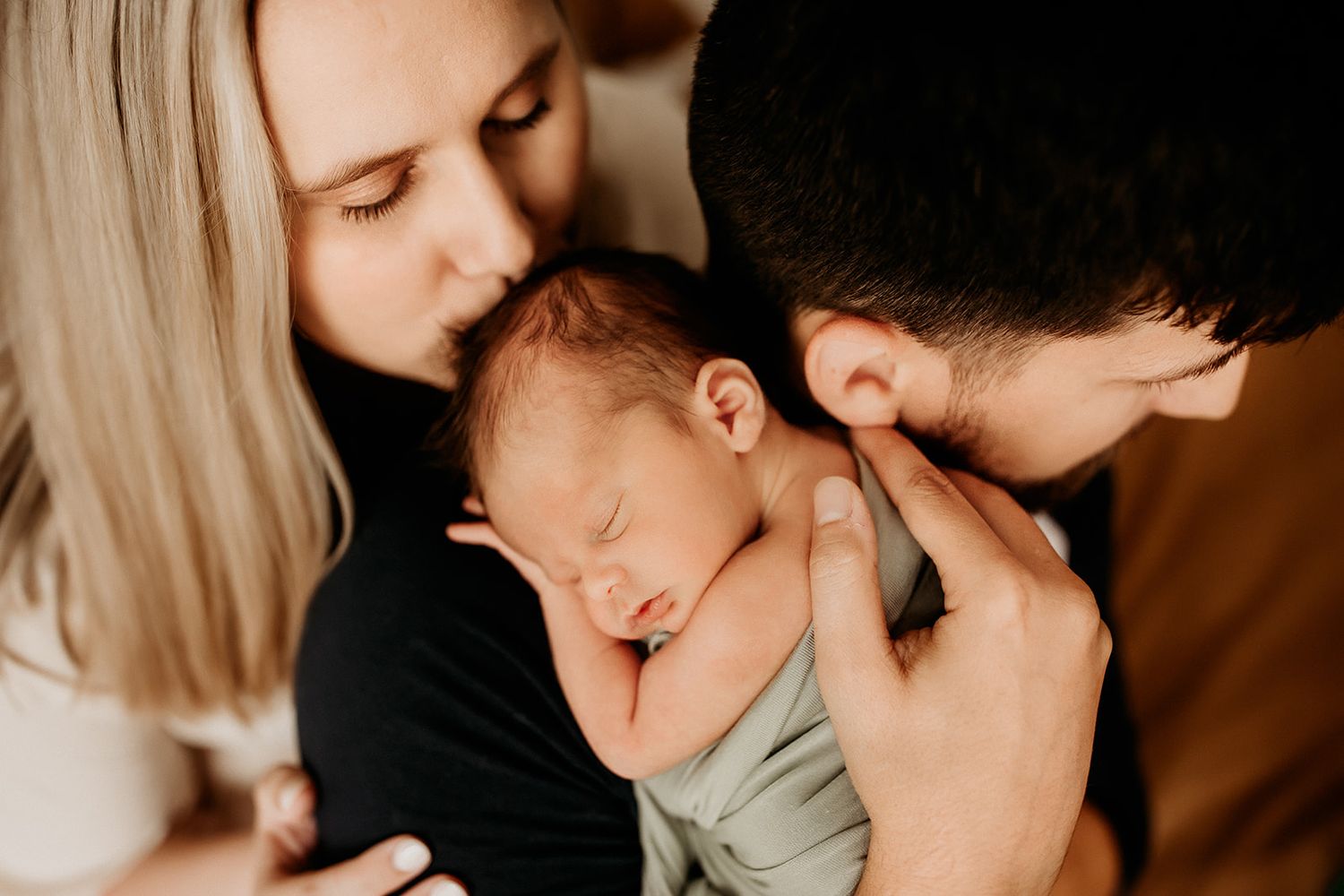 A father holds his baby boy on his shoulder while the mother leans over and kisses the top of his head.