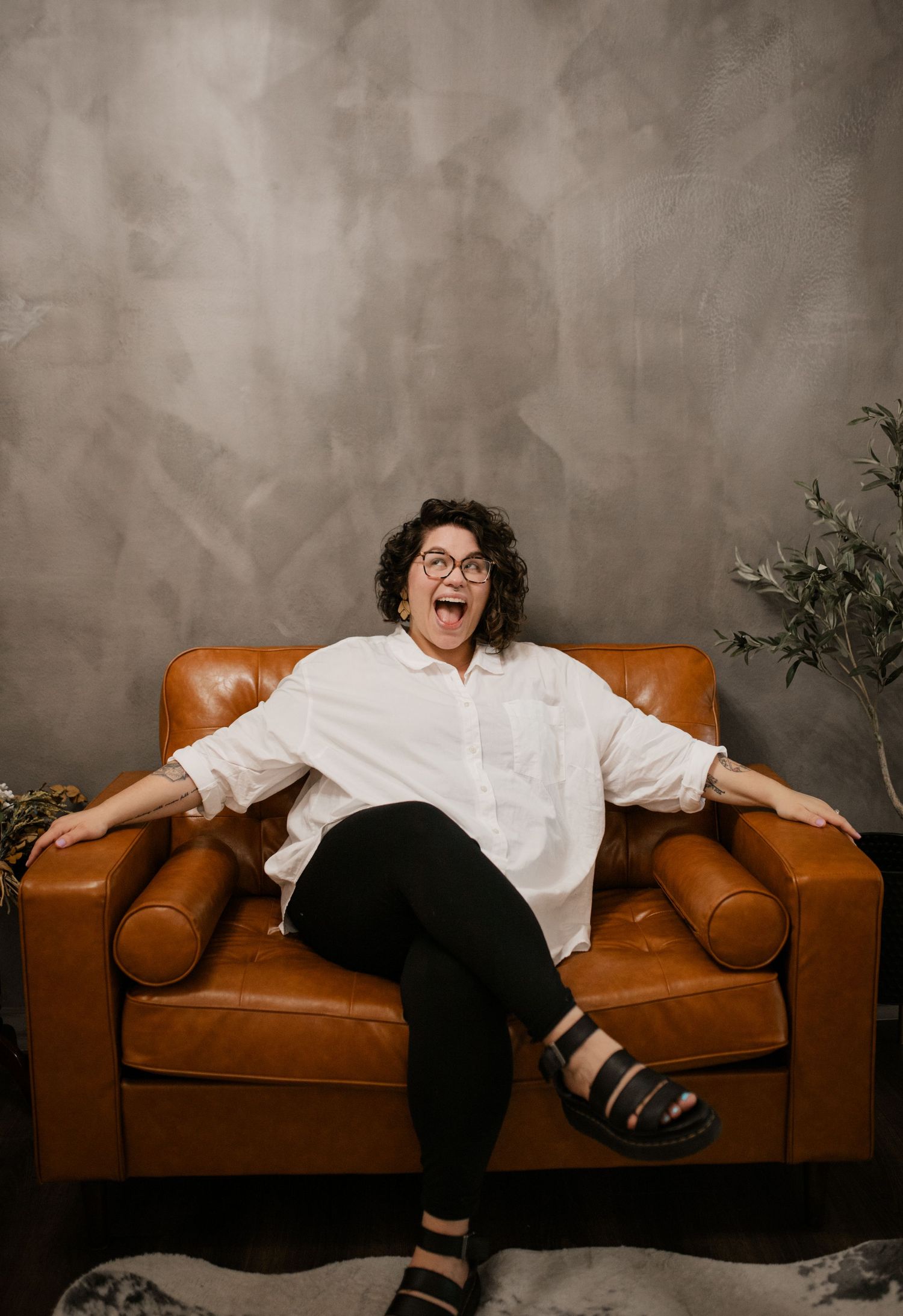 A person in white shirt and black pants poses expressively on a brown leather chair.