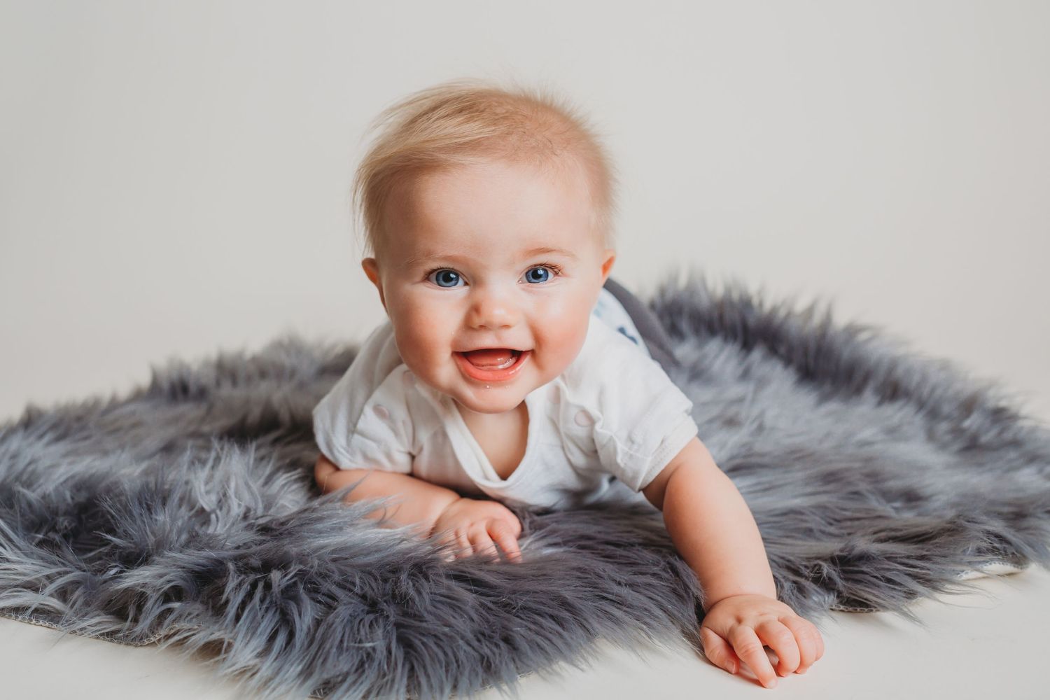 Smiling baby lying on soft gray fur rug during studio photography session.