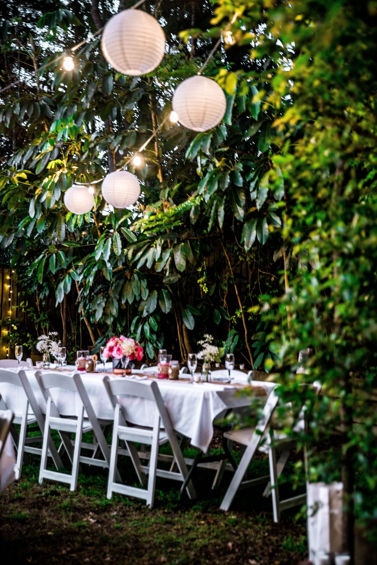 White paper lanterns illuminate an outdoor dining setup against lush green foliage.