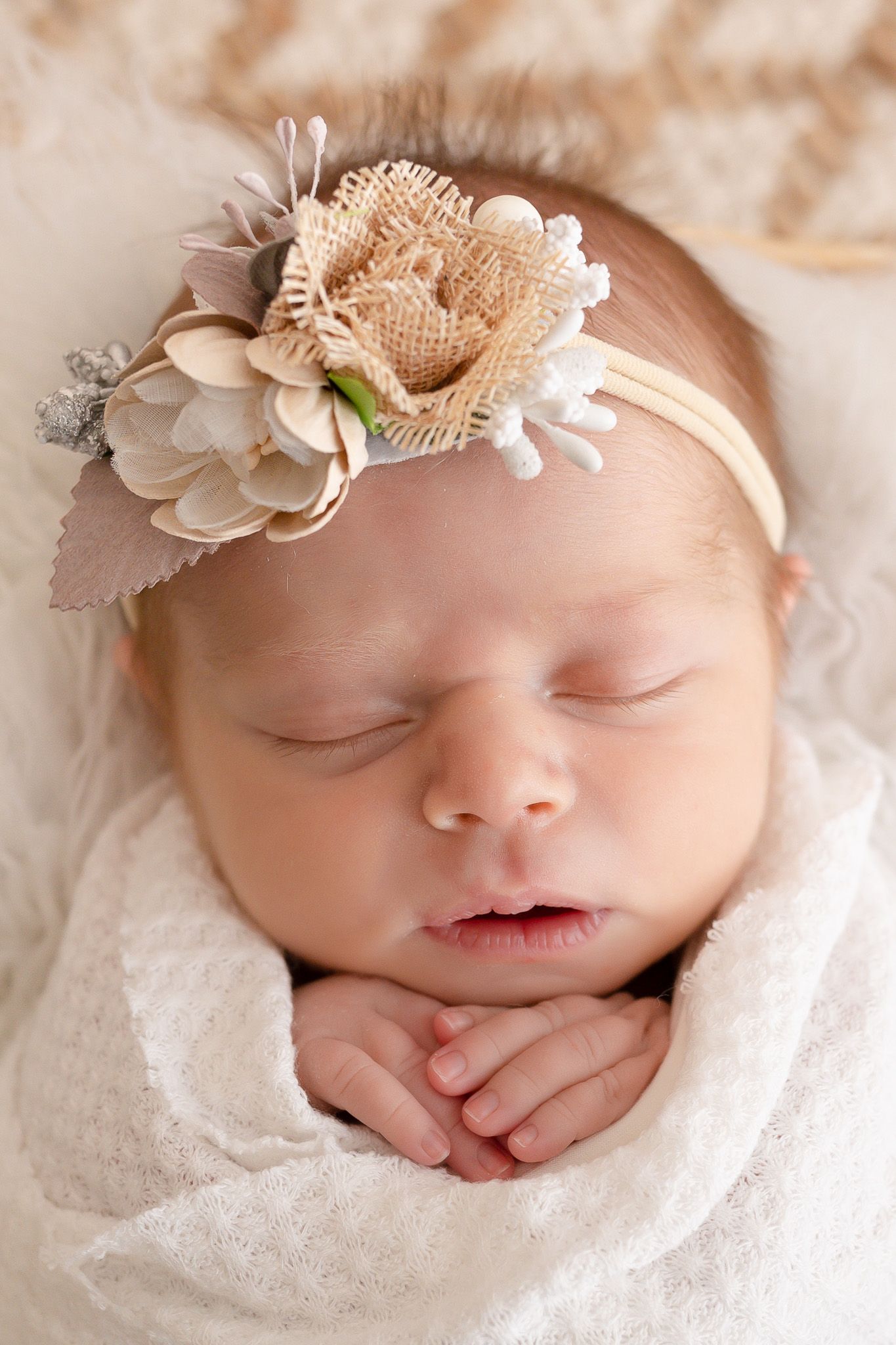 Sleeping newborn wearing delicate beige floral headband wrapped in soft white blanket.