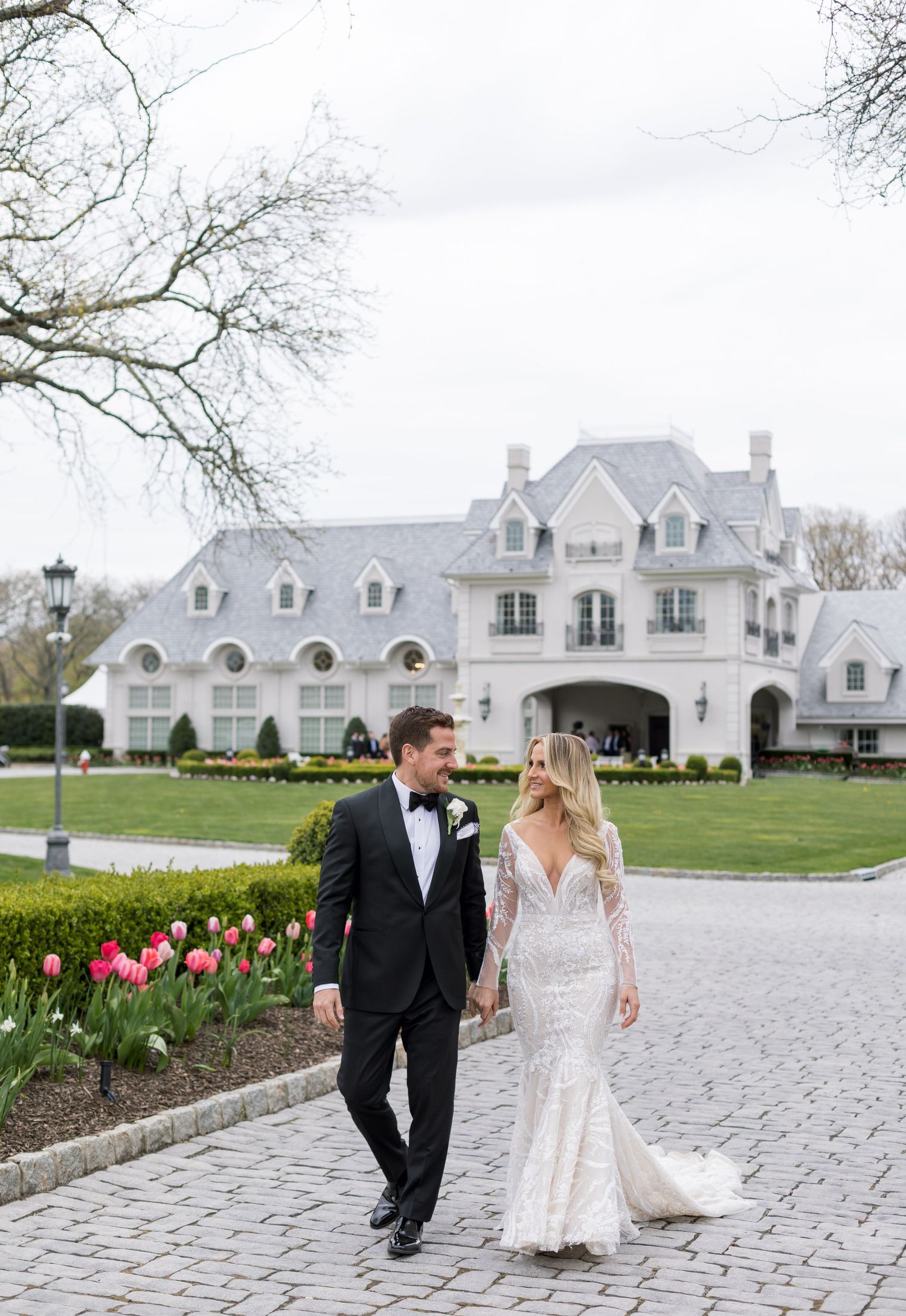 Couple takes romantic stroll along pathway by grand white mansion with blooming spring tulips.
