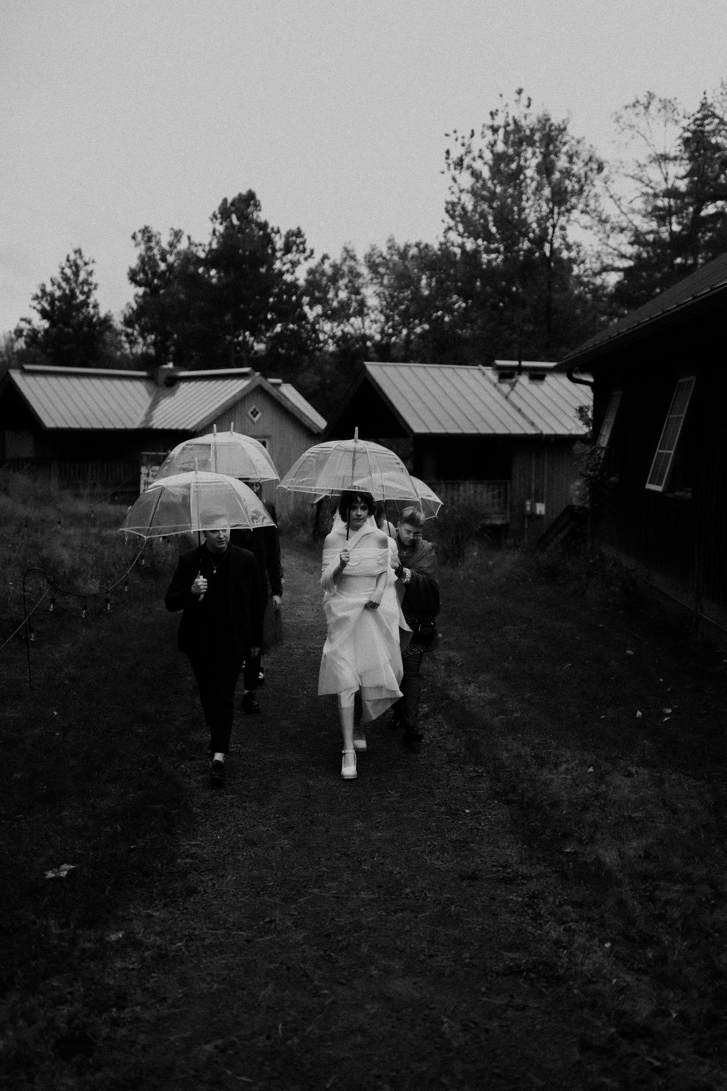 A black and white photo of people walking with umbrellas near rustic cabin buildings.