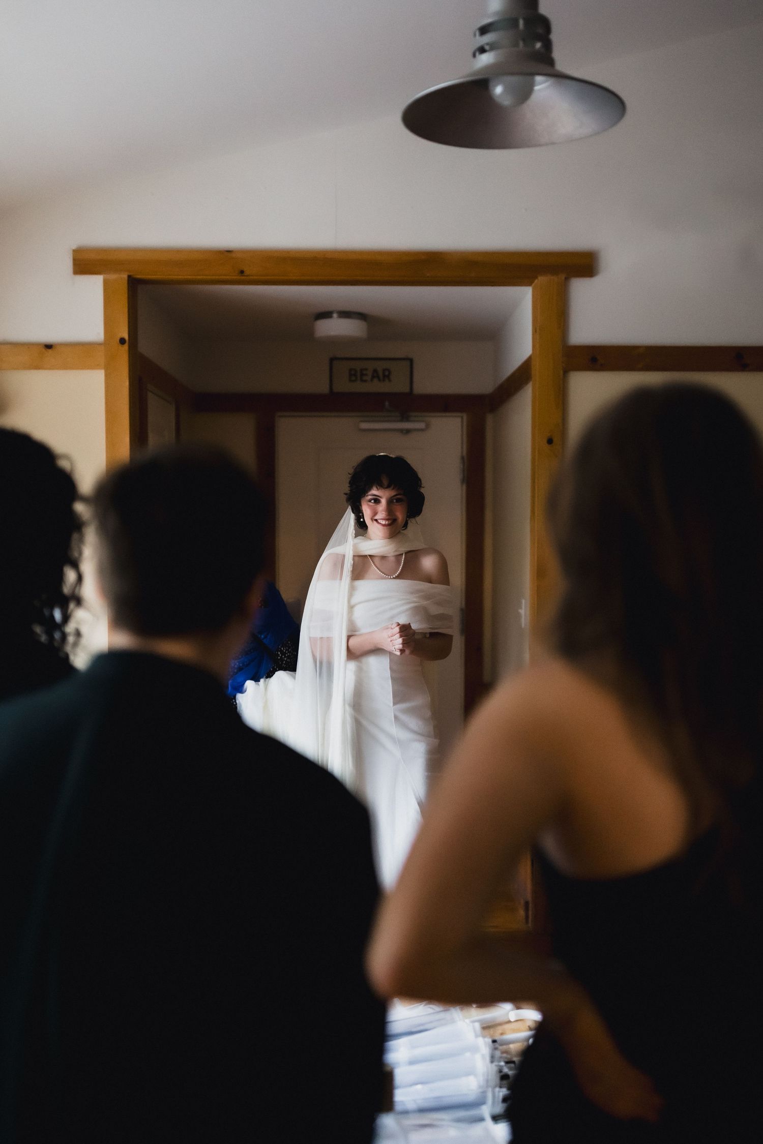 A bride in white stands in a doorway as others watch during a wedding preparation moment.