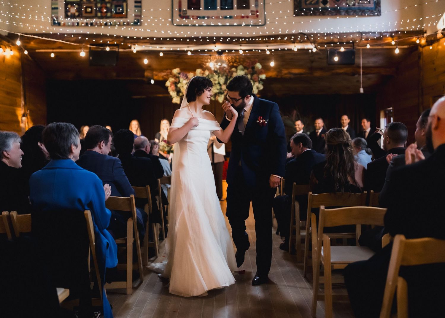 A wedding ceremony in a rustic venue decorated with string lights as guests watch the couple's first dance.