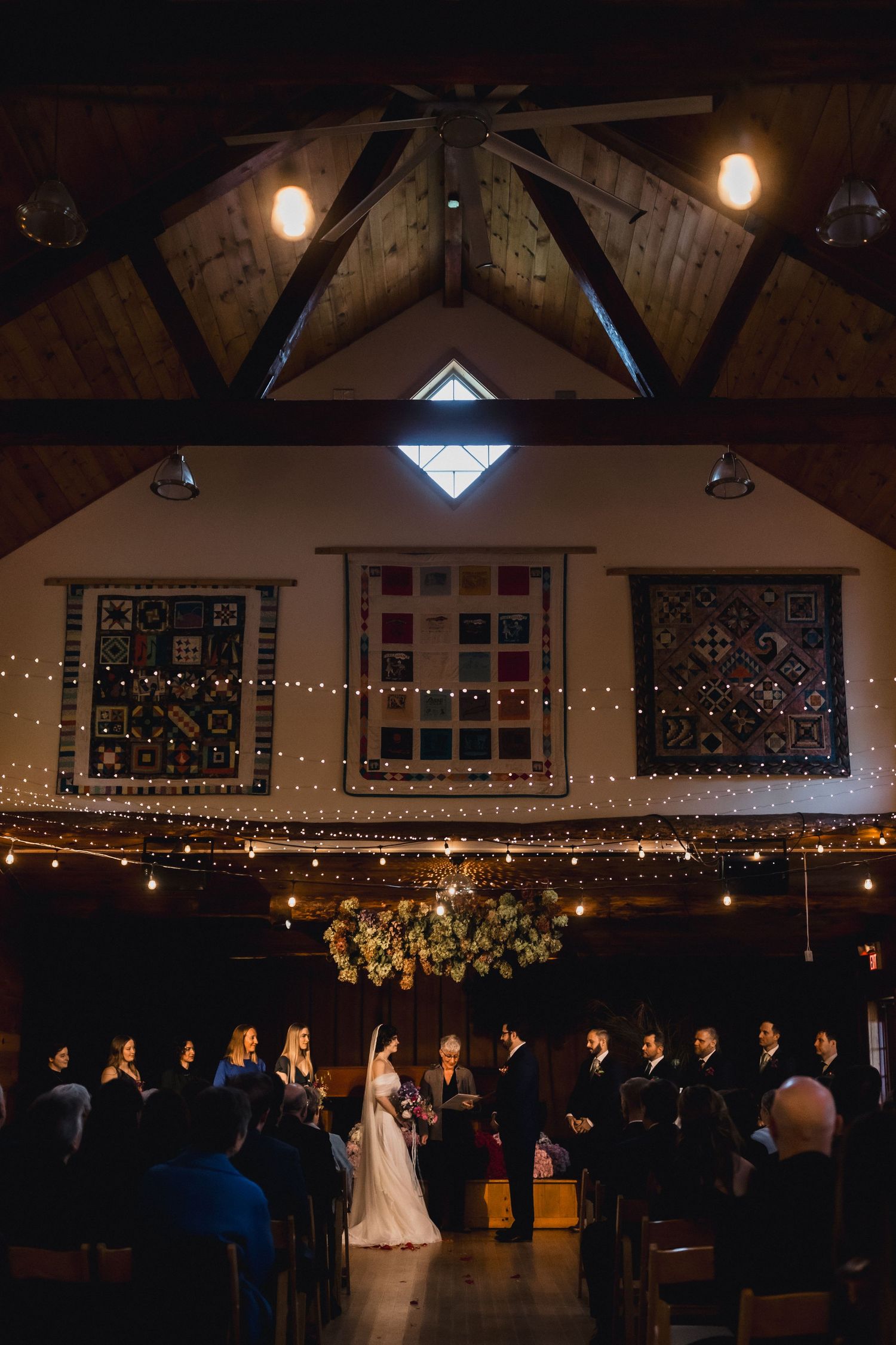 Wedding ceremony in rustic barn with exposed beams, string lights and decorative quilts on walls.