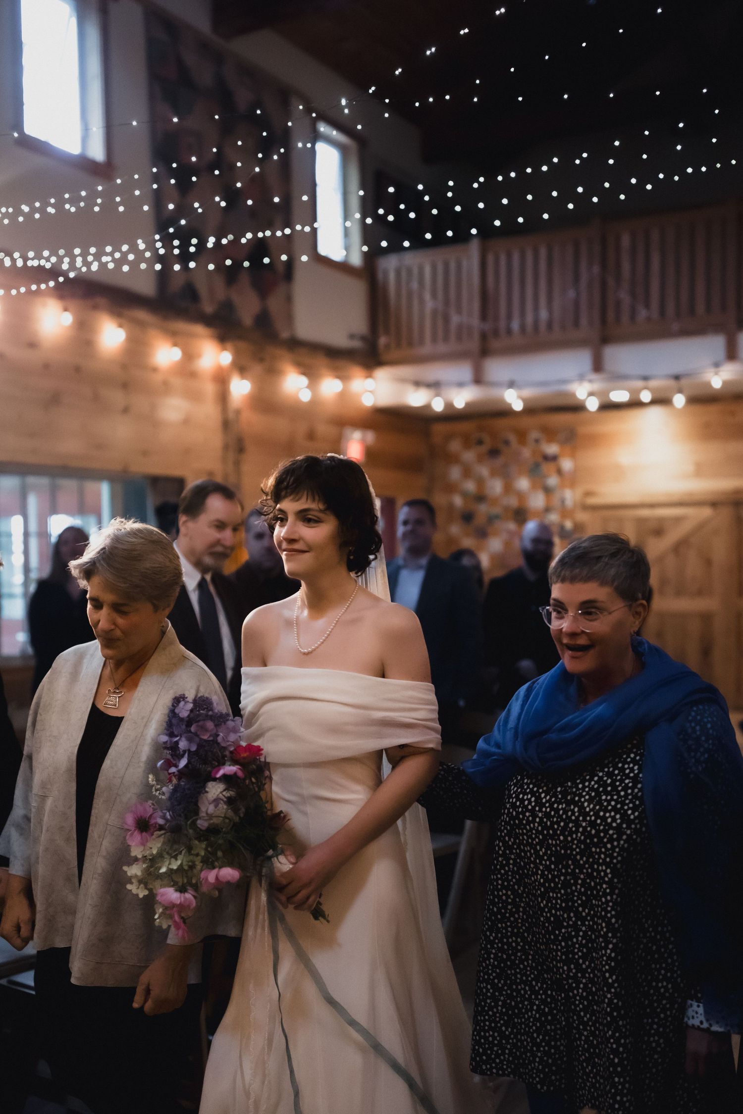 Wedding celebration with guests in elegant attire gathering under twinkling string lights in rustic venue.