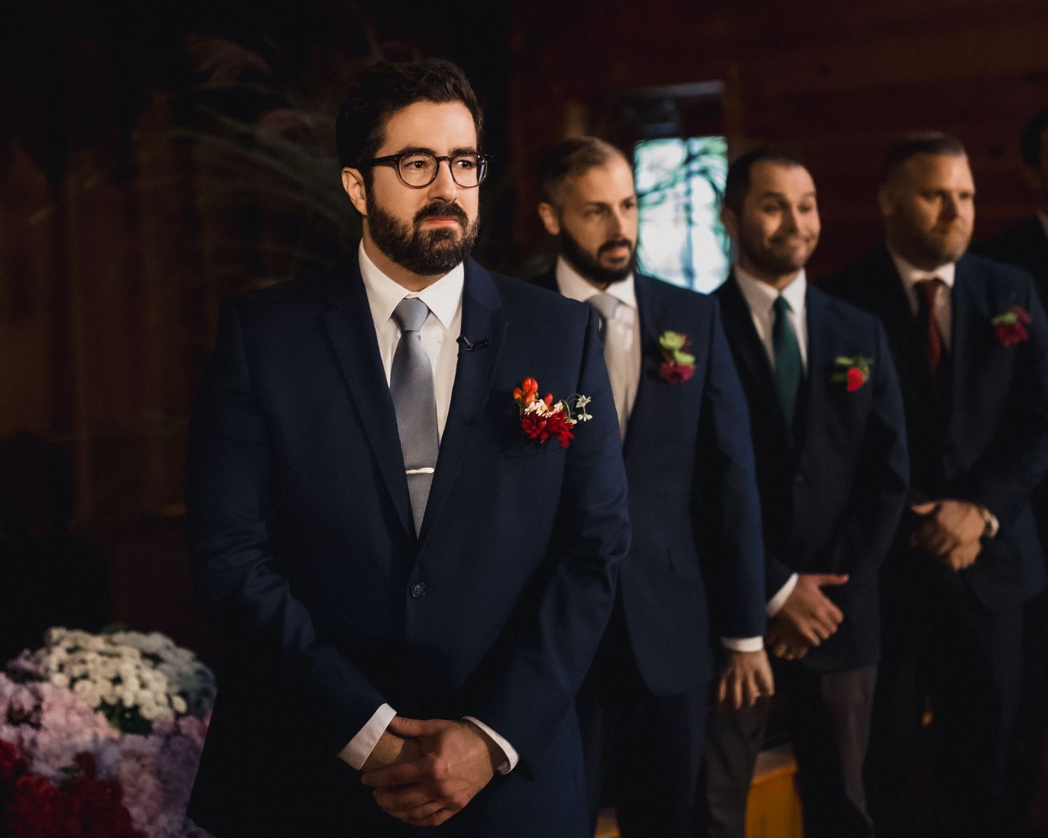 Wedding party groomsmen in navy suits with red boutonnieres stand in formation at rustic indoor venue.