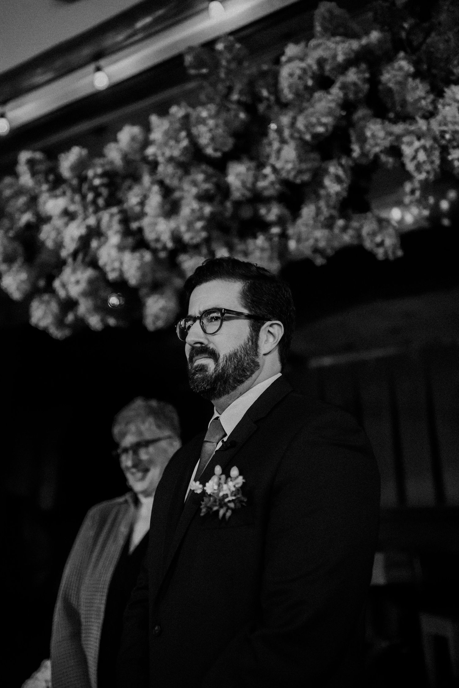 A groom stands beneath floral decorations during a wedding ceremony in black and white.