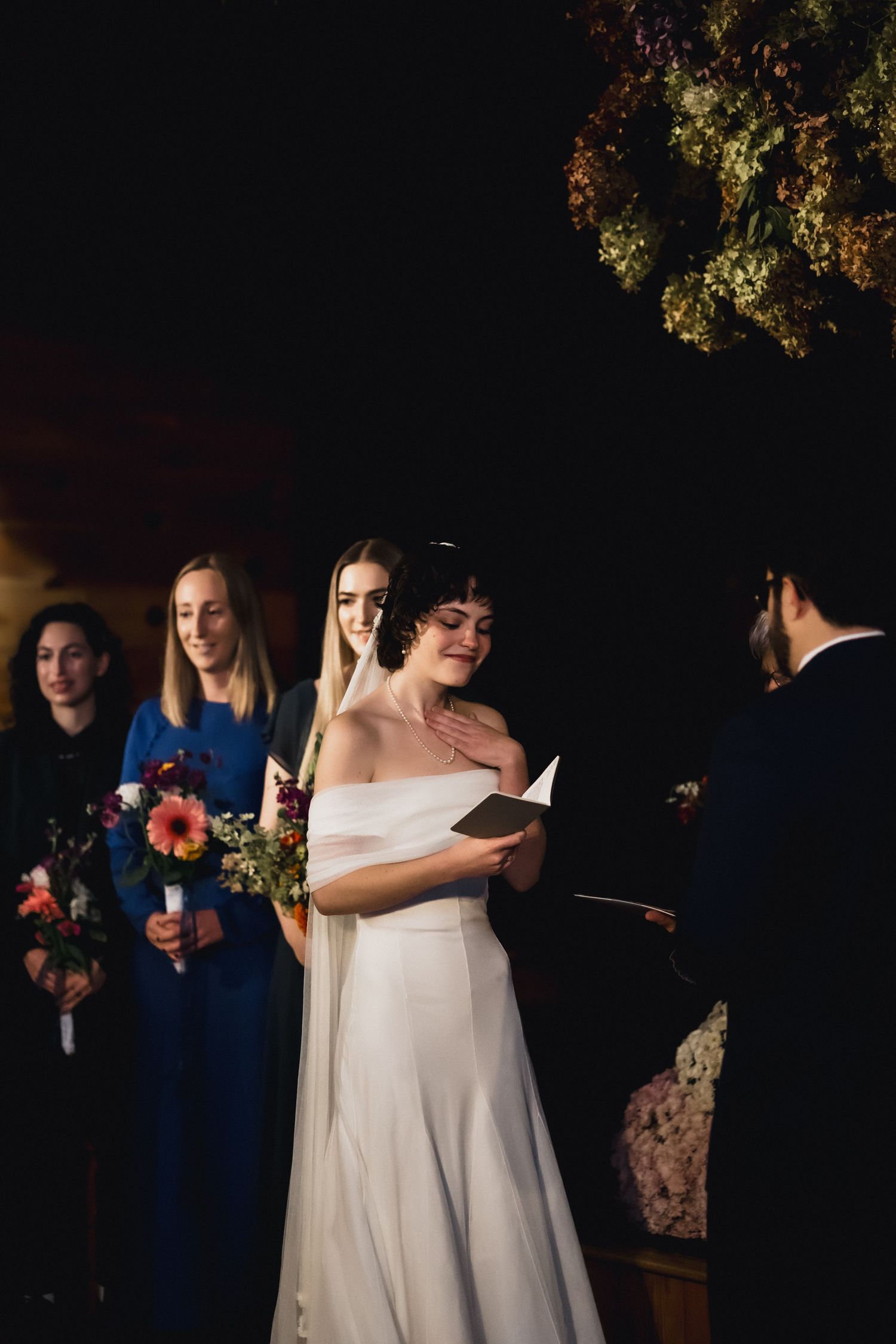 Wedding party during ceremony under floral arrangements in rustic indoor venue.