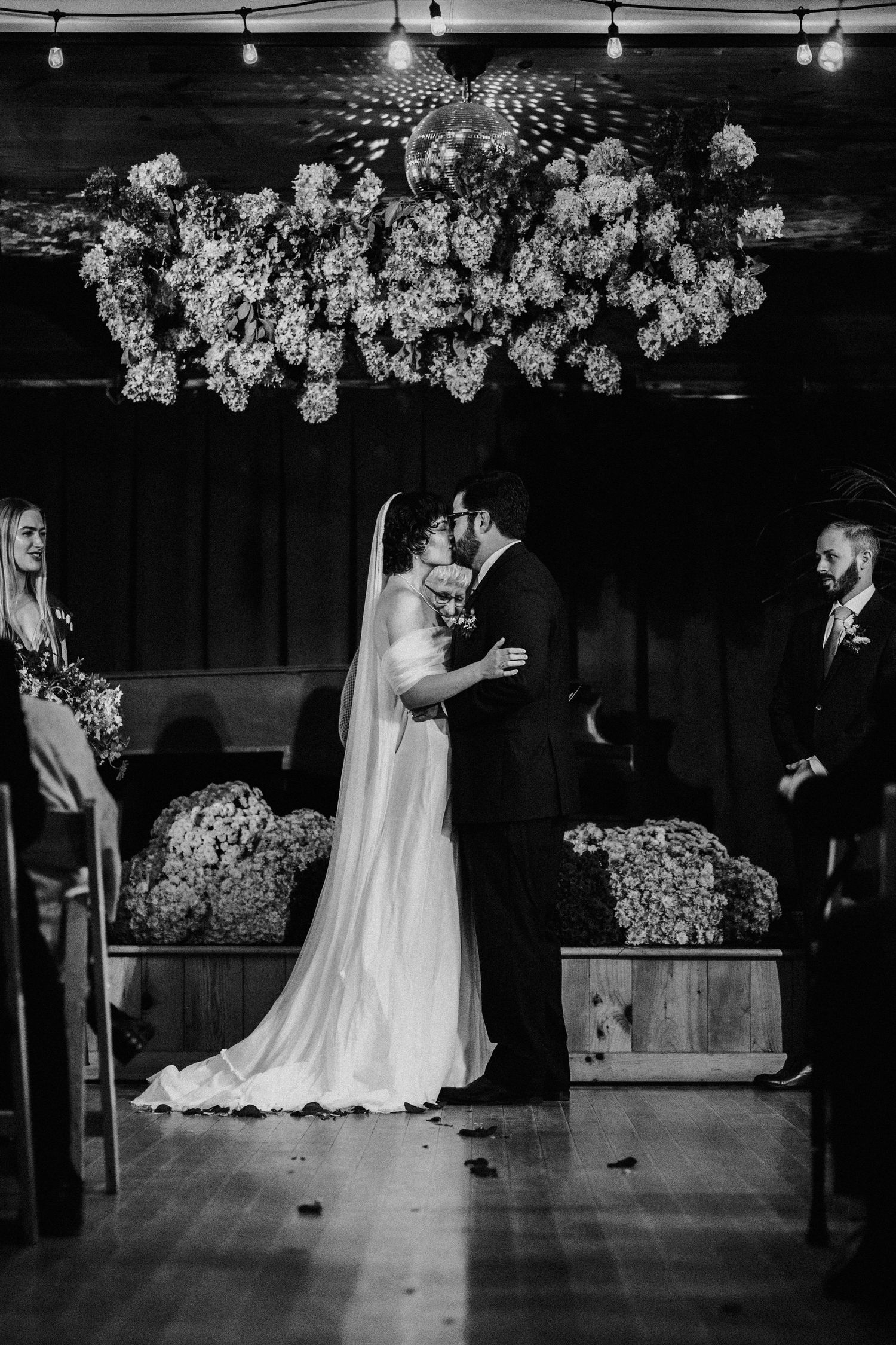 Black and white wedding ceremony photo under dramatic floral chandelier in rustic venue.