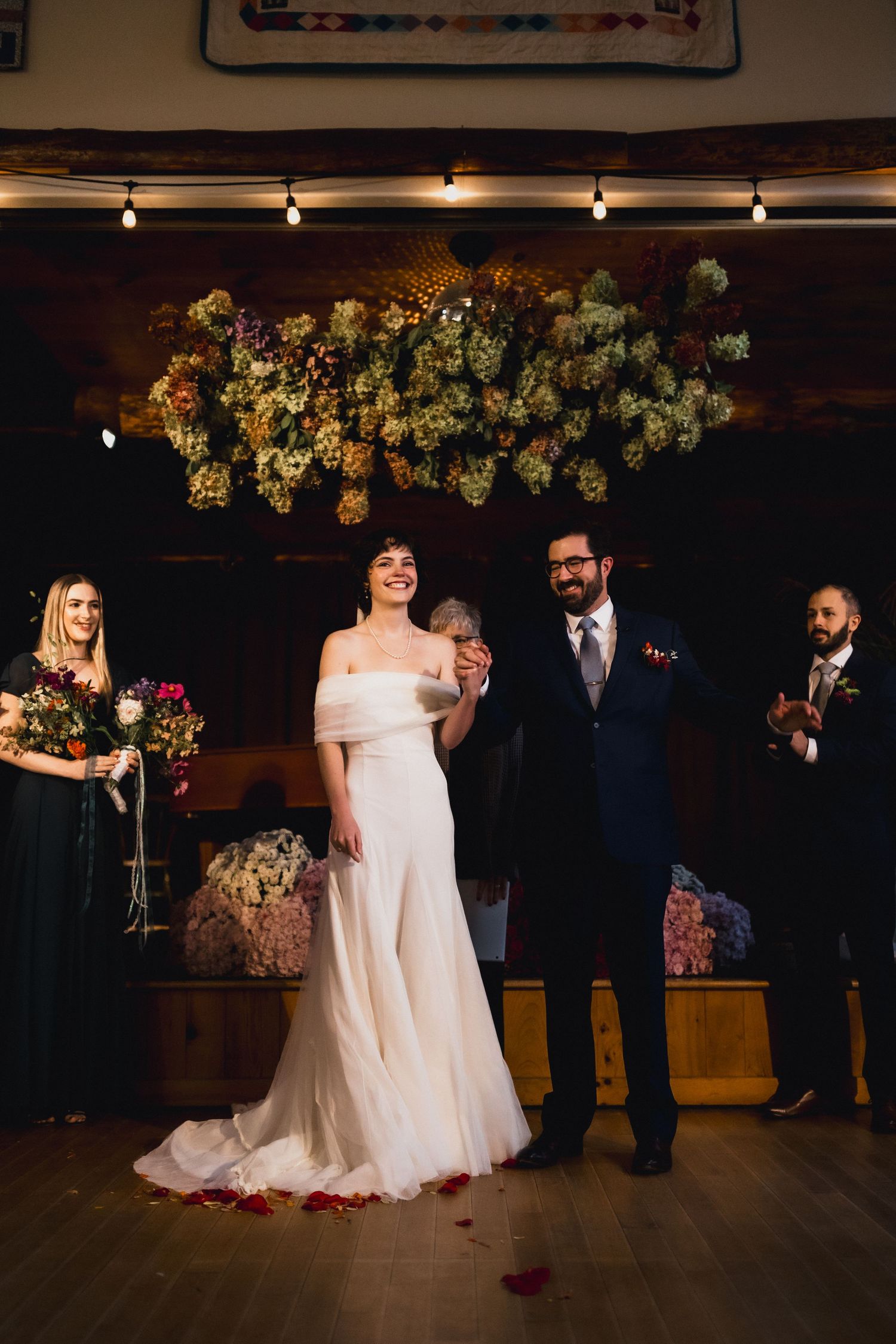 Wedding couple stands under suspended floral arrangement during ceremony in rustic venue.