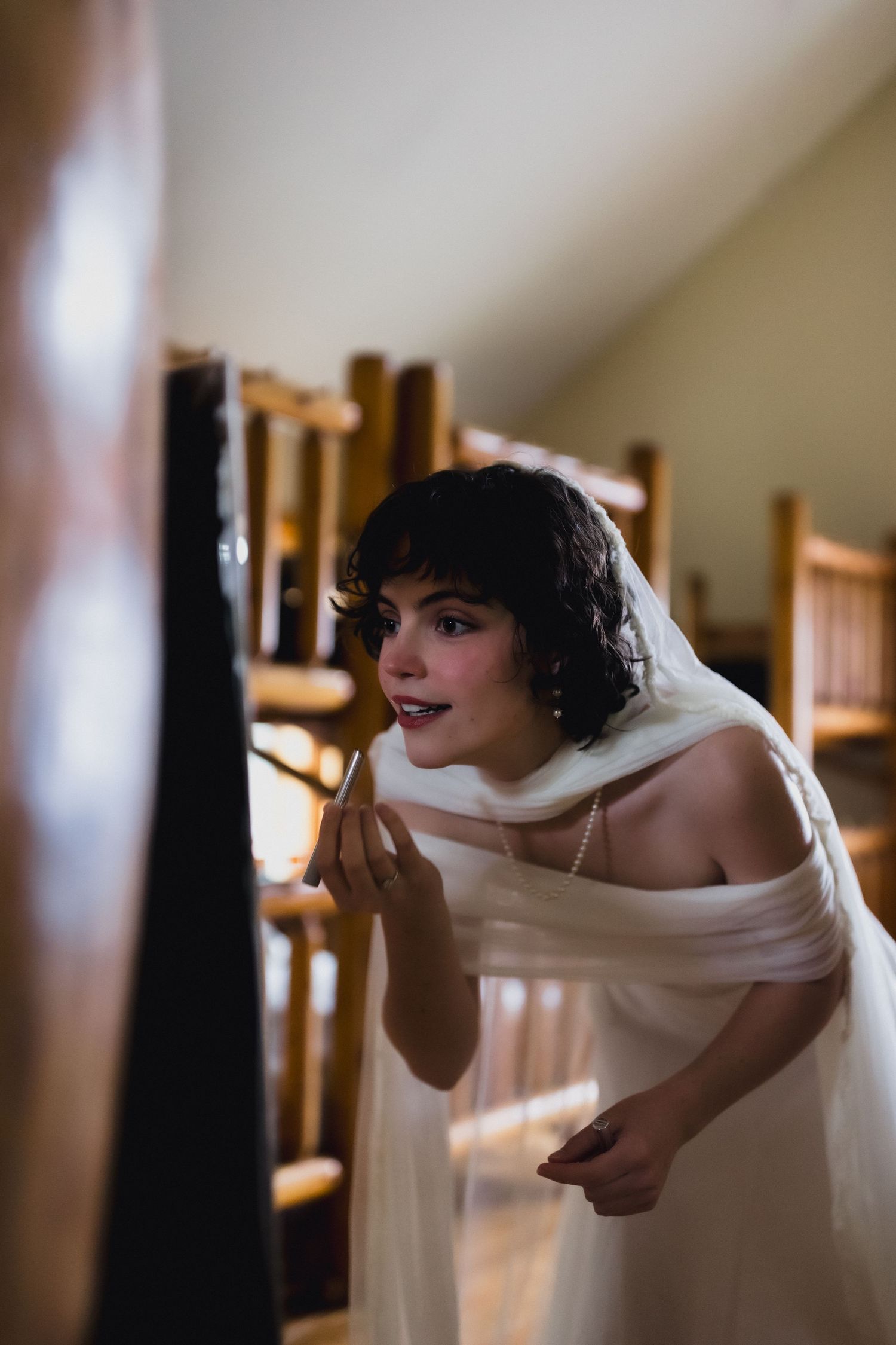 A bride in a white off-shoulder dress applies makeup while looking in a mirror.