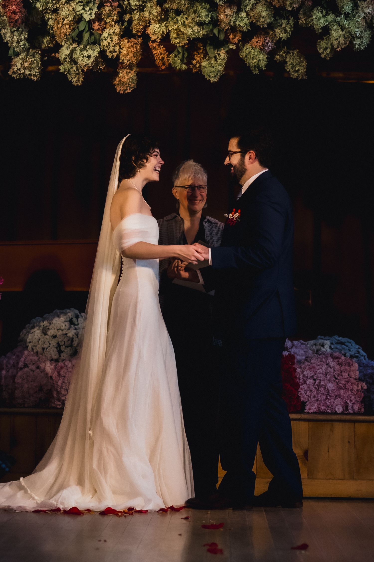Wedding ceremony exchange of rings under floral decor in rustic venue with warm lighting.